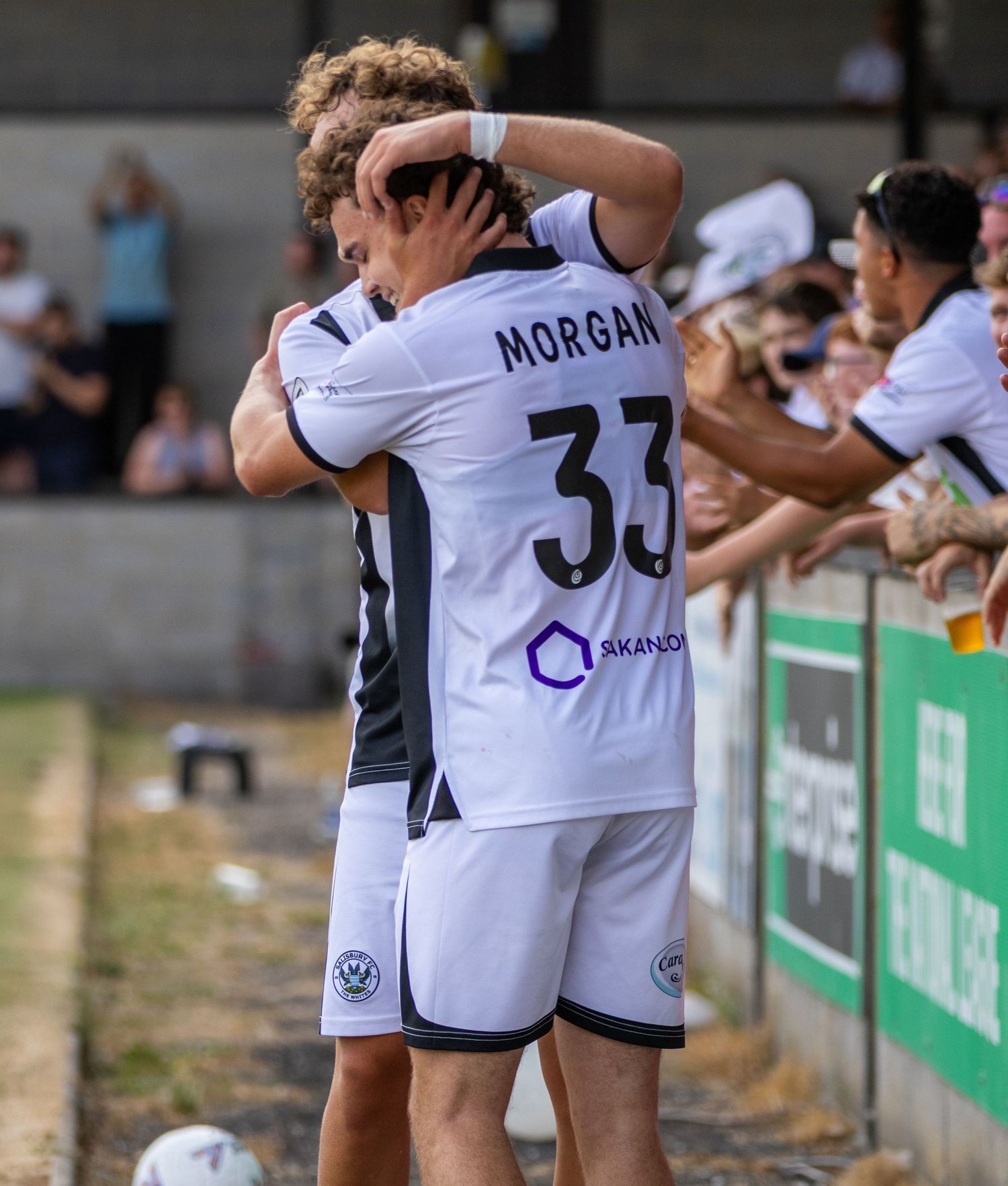 Two footballers in white kits embrace near the touchline as fans celebrate, with player number 33 Morgan visible.