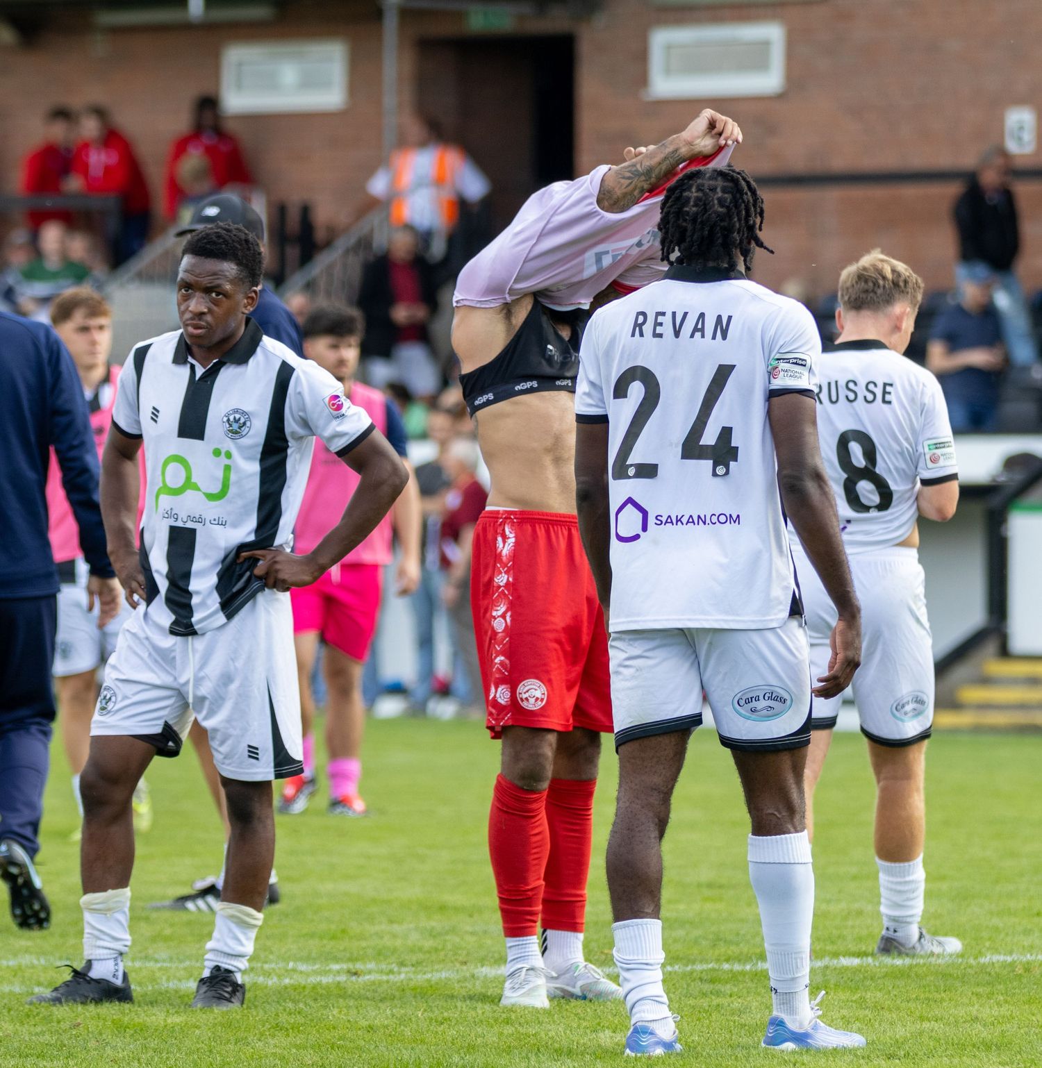 Football players in black and white jerseys, including number 24 Revan, gather on a pitch after a match.