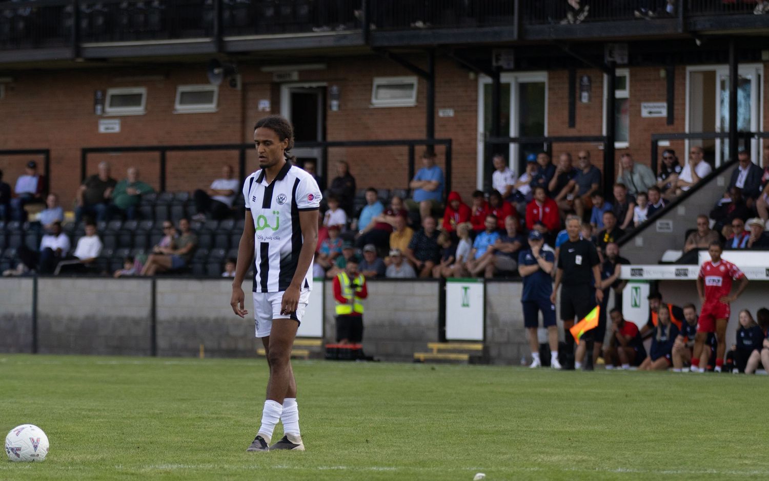 A footballer in black and white kit stands near the ball during a match at a small stadium with spectators in the stands.