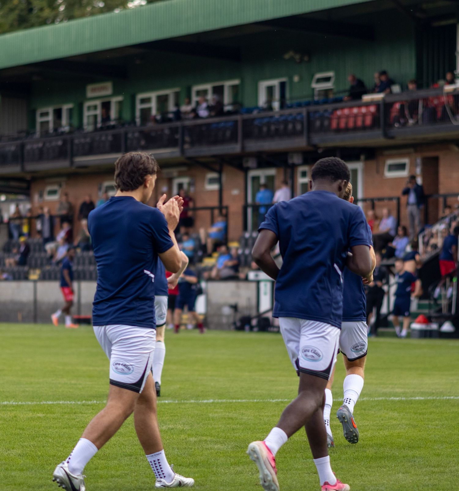 Two footballers in navy blue kits warm up by jogging on a grass pitch with a crowded stand behind them.