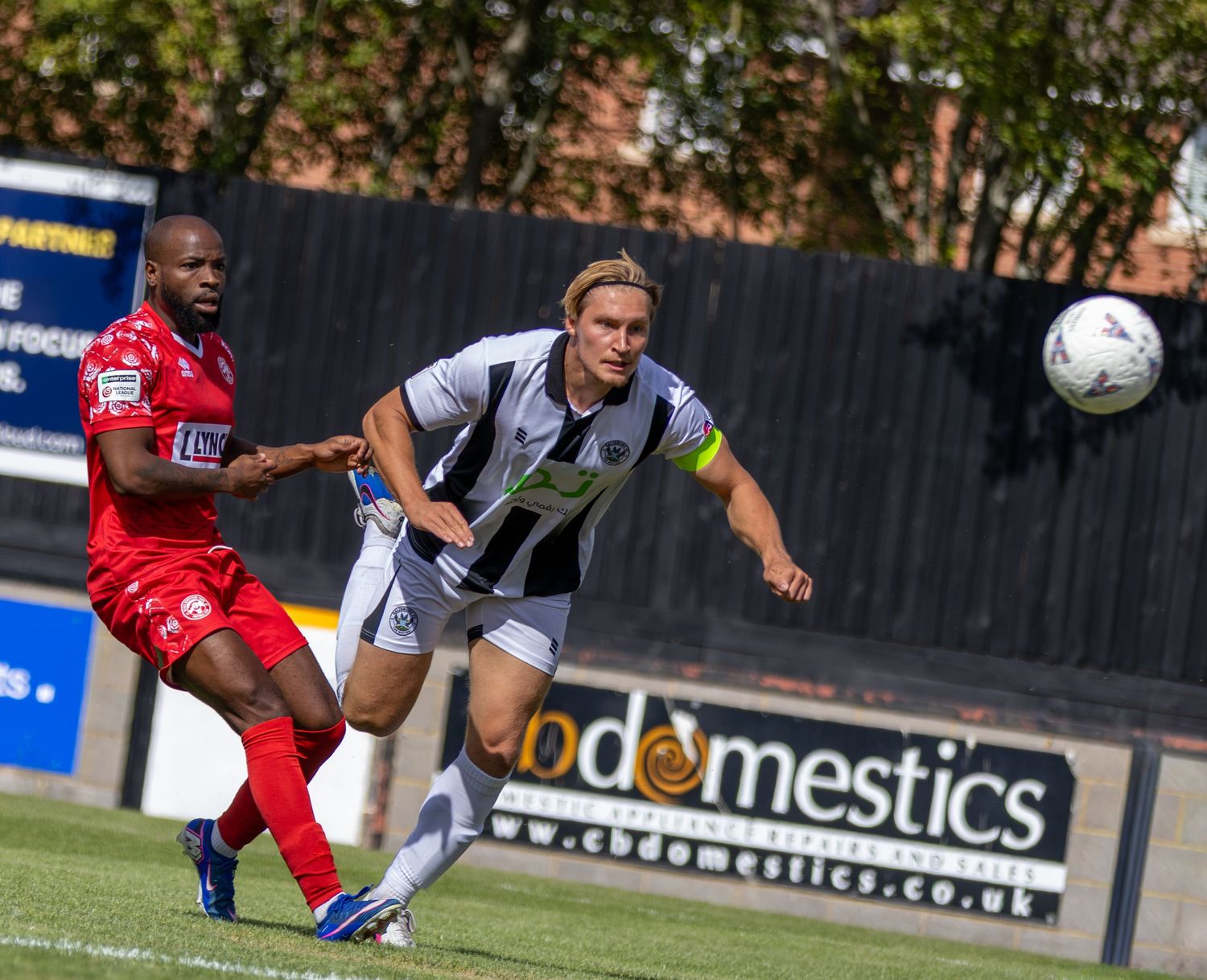 Two soccer players compete for the ball during a match, one in red and one in black and white kit.