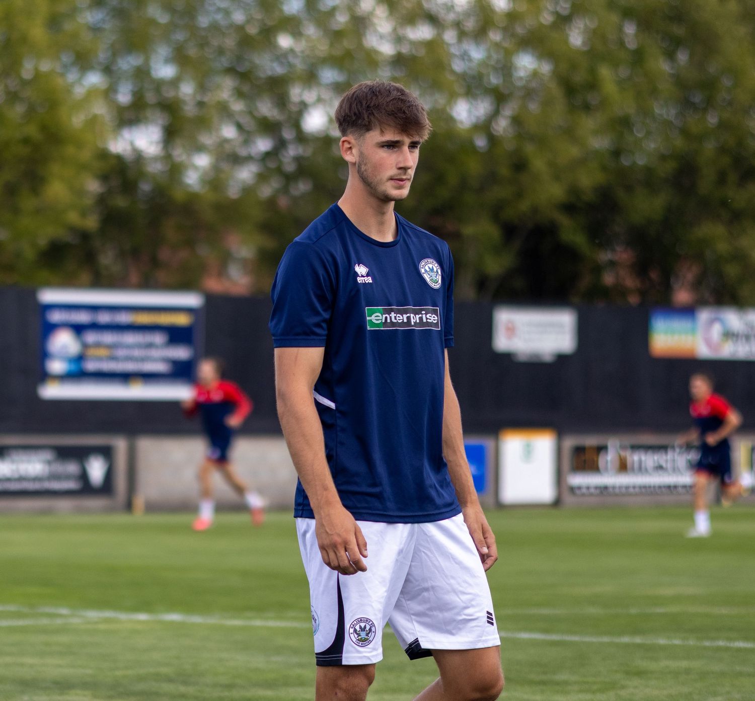 Young male footballer in navy enterprise-sponsored Errea kit stands on a grass pitch during pre-match warmup.