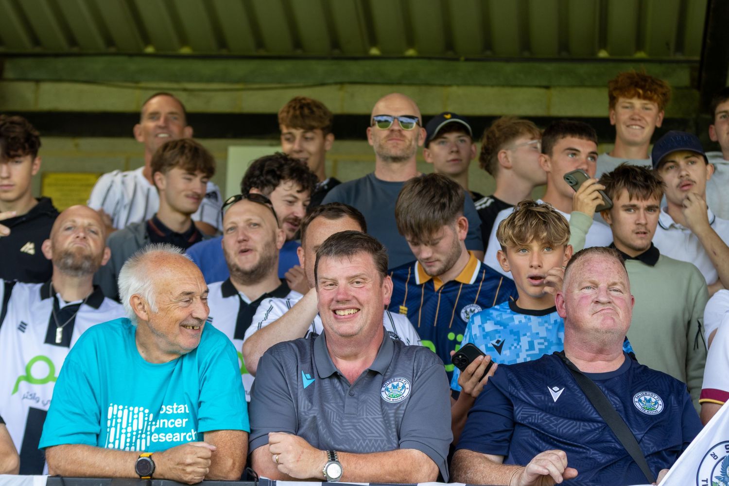 Crowd of football fans smiling in stadium stands on sunny matchday including older and younger supporters.