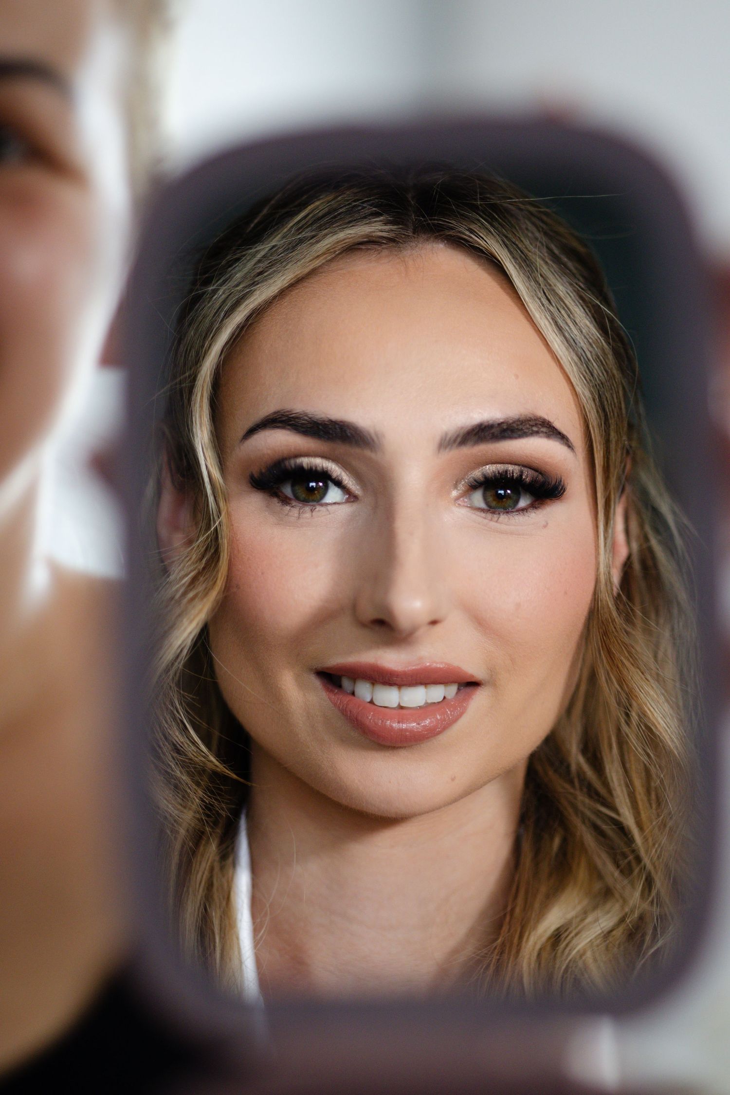 Smiling blonde woman with stunning bridal makeup reflected in a handheld mirror with soft lighting.