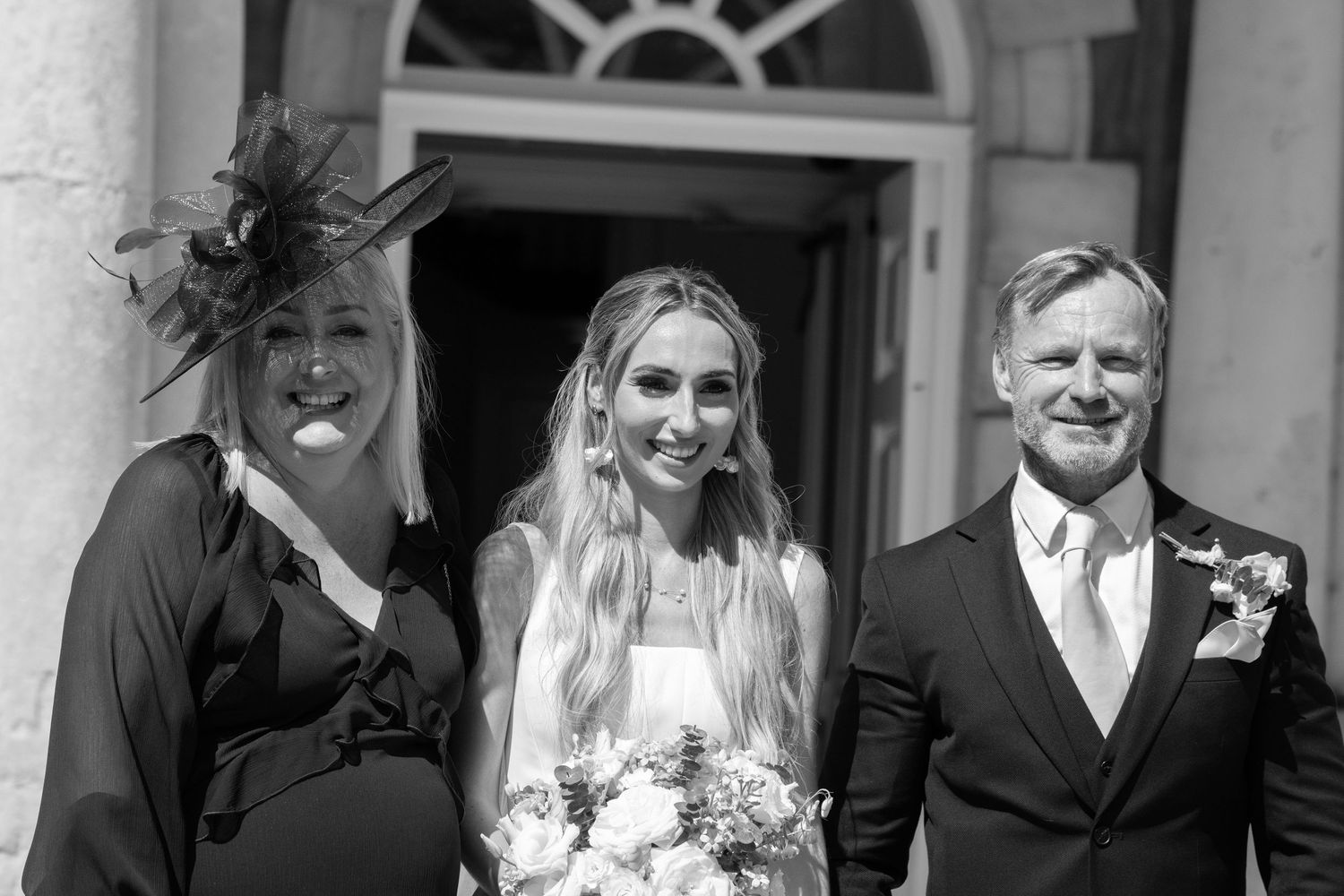 Black and white wedding photo of smiling bride with bouquet between woman in feathered hat and suited man.
