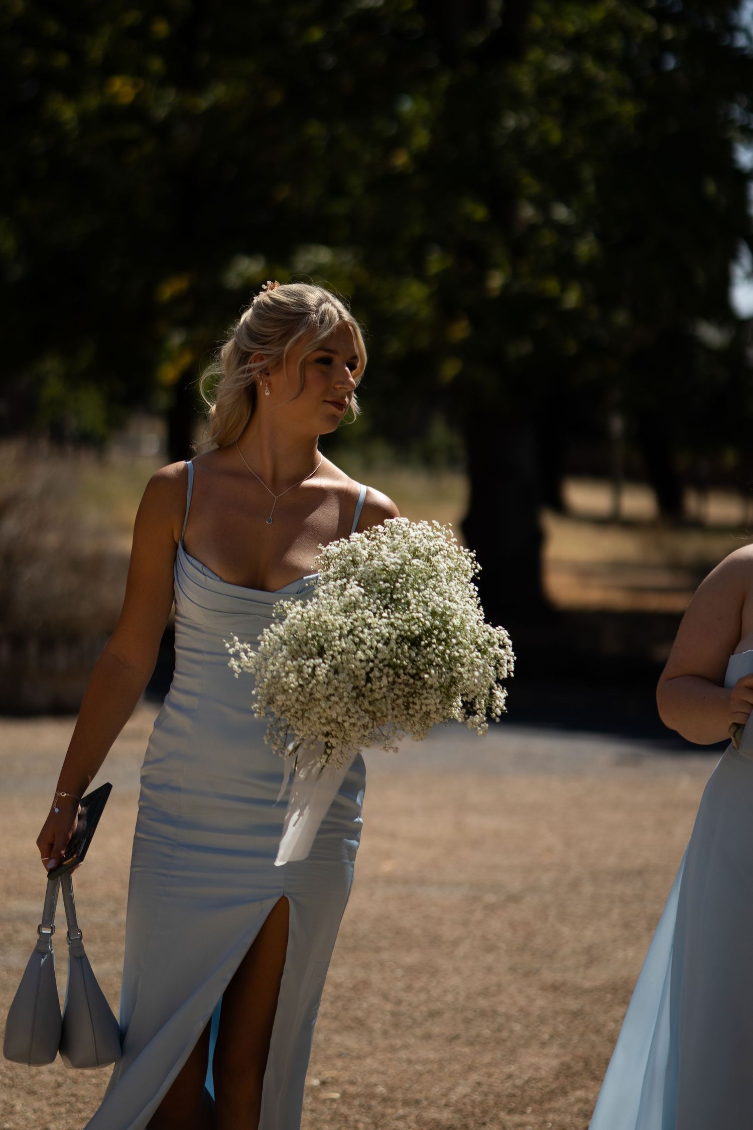 A bridesmaid in a light blue satin dress carries a bouquet of white gypsophila flowers outdoors.