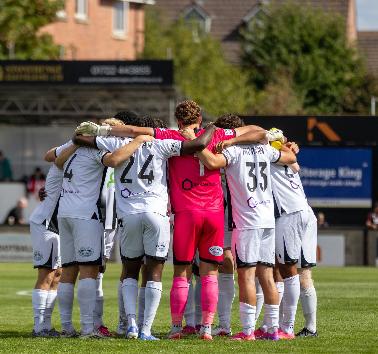 Soccer team forms pre-match huddle on green pitch wearing white uniforms with pink-kitted goalkeeper.