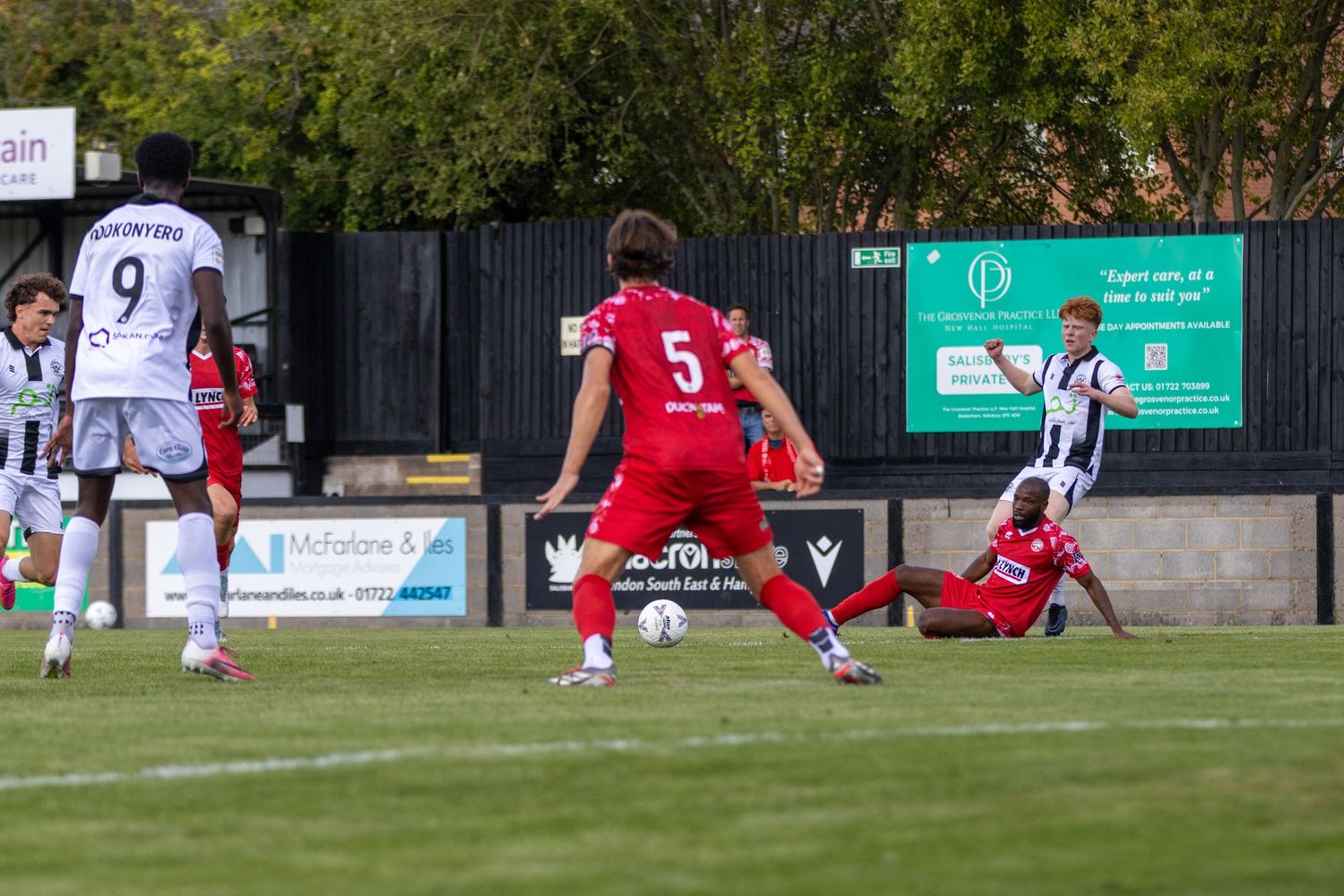 Non-league football match action as red and white kit players compete for the ball on a green pitch.