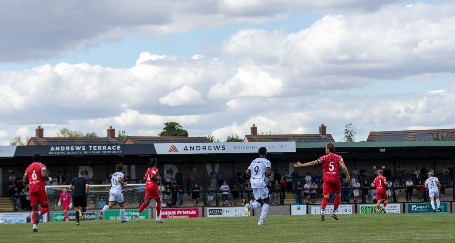 Football match in progress at Andrews Terrace ground with players in red and white kits competing on a sunny day.