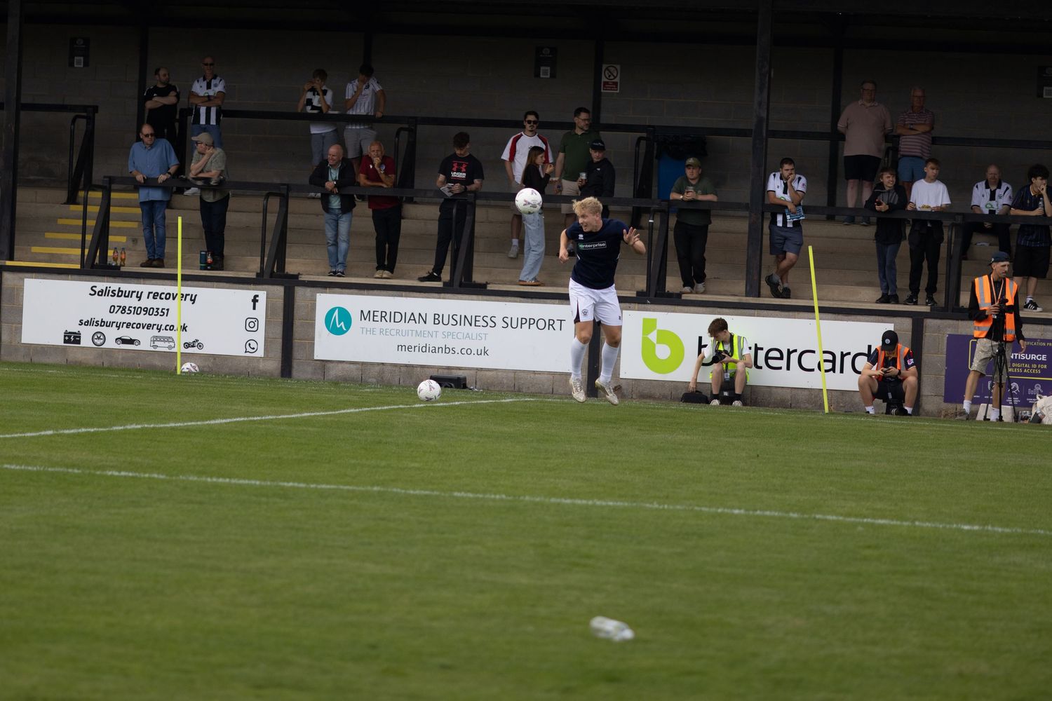 A footballer in dark kit prepares to take a free kick on a grass pitch with spectators watching.