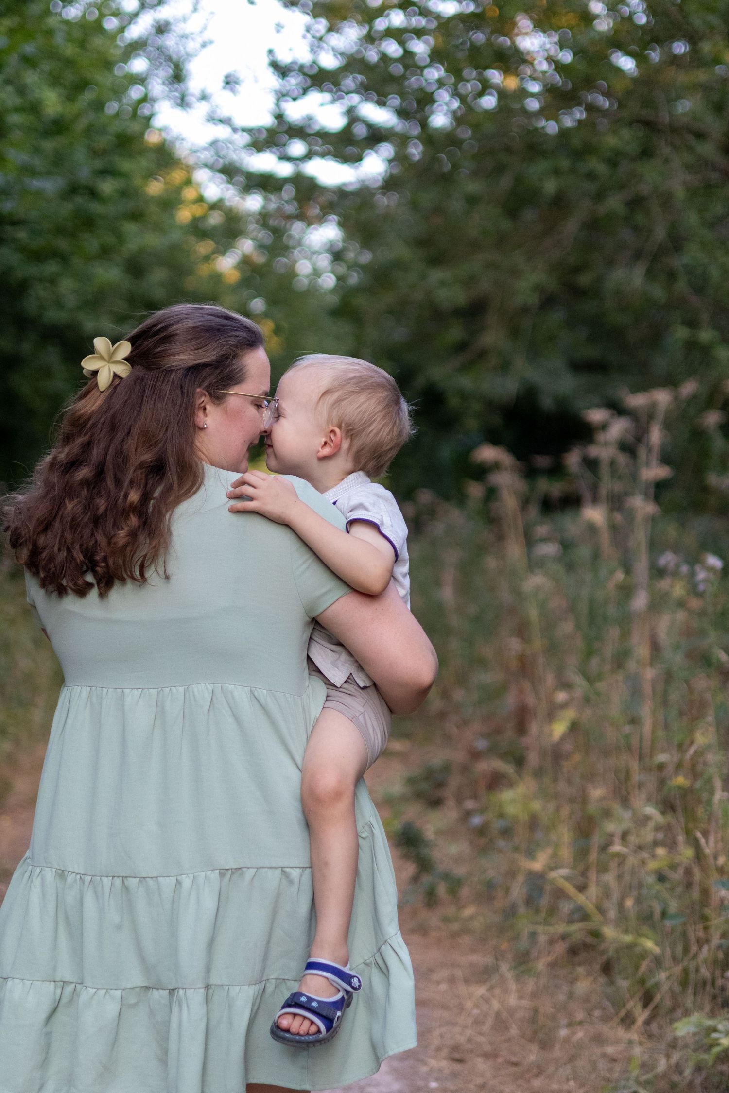 A loving mother in a mint green dress shares an Eskimo kiss with her baby on a forest path.