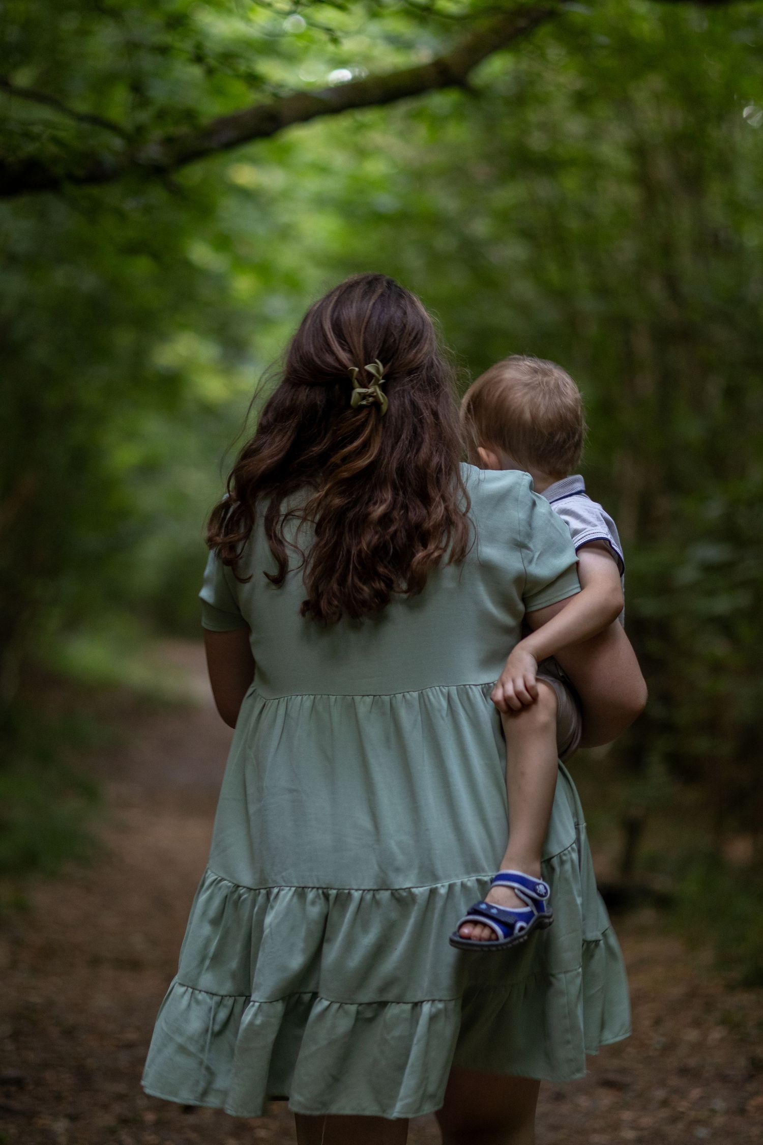 Back view of a brunette woman in mint dress carrying a young child on a woodland trail.