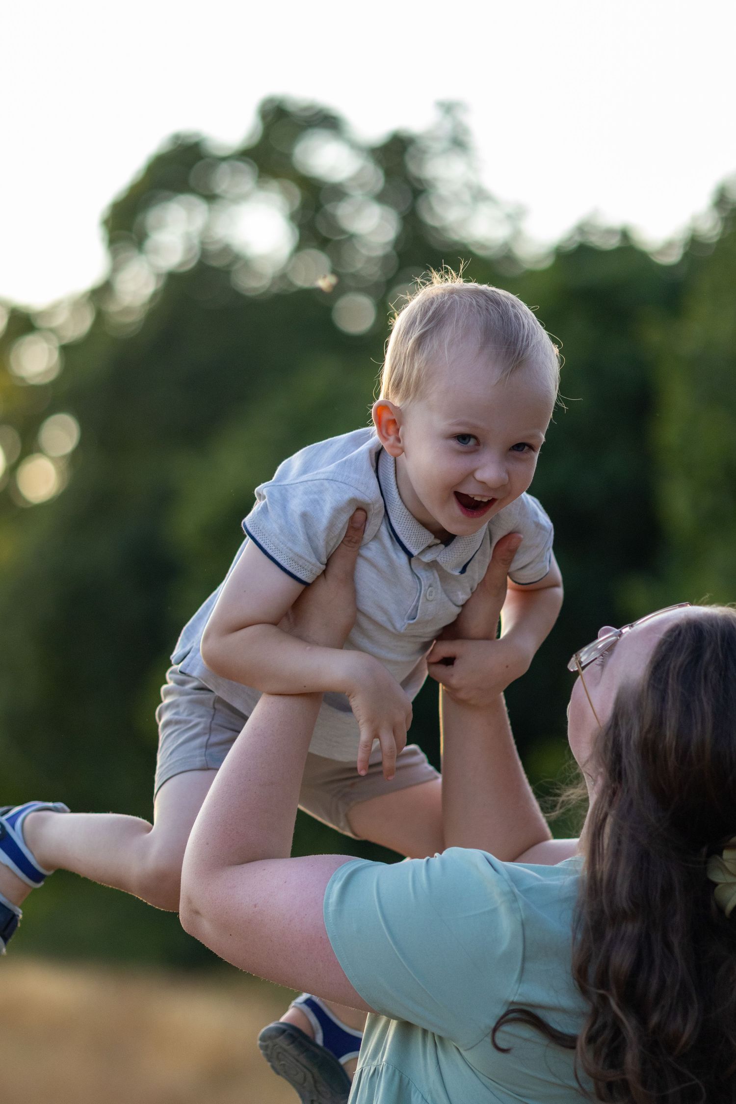 Joyful toddler laughing while being lifted in the air by mother outdoors at sunset.