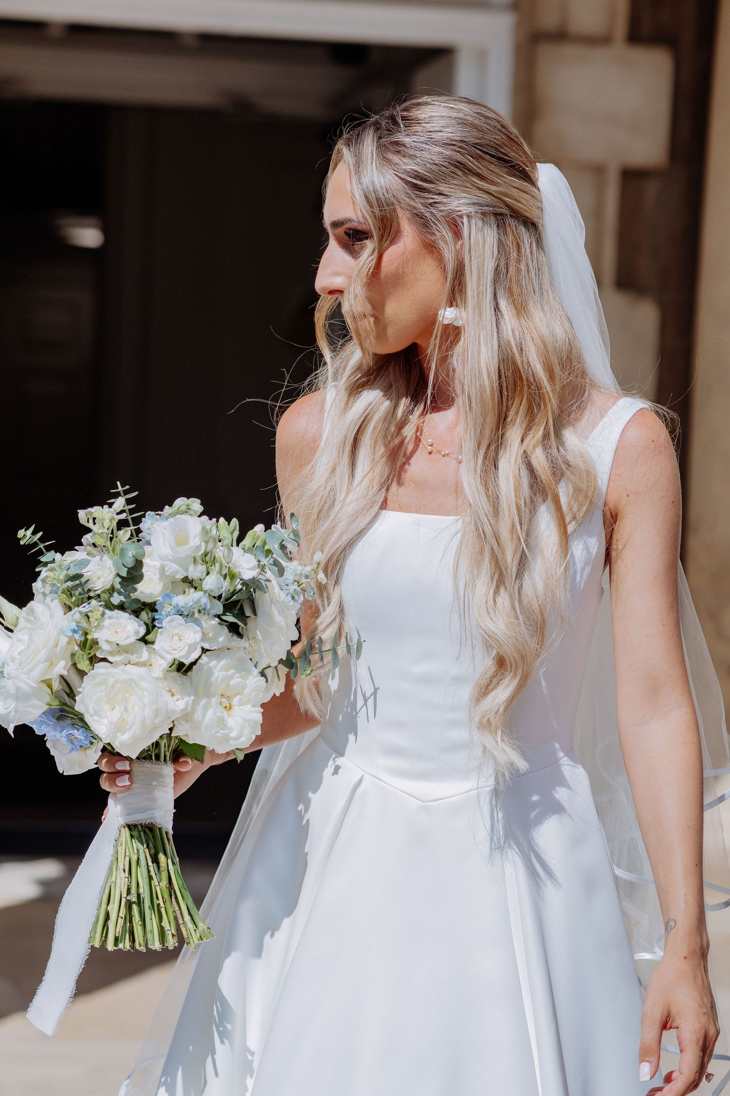 Beautiful blonde bride in white wedding dress and veil holds elegant white floral bouquet outdoors.
