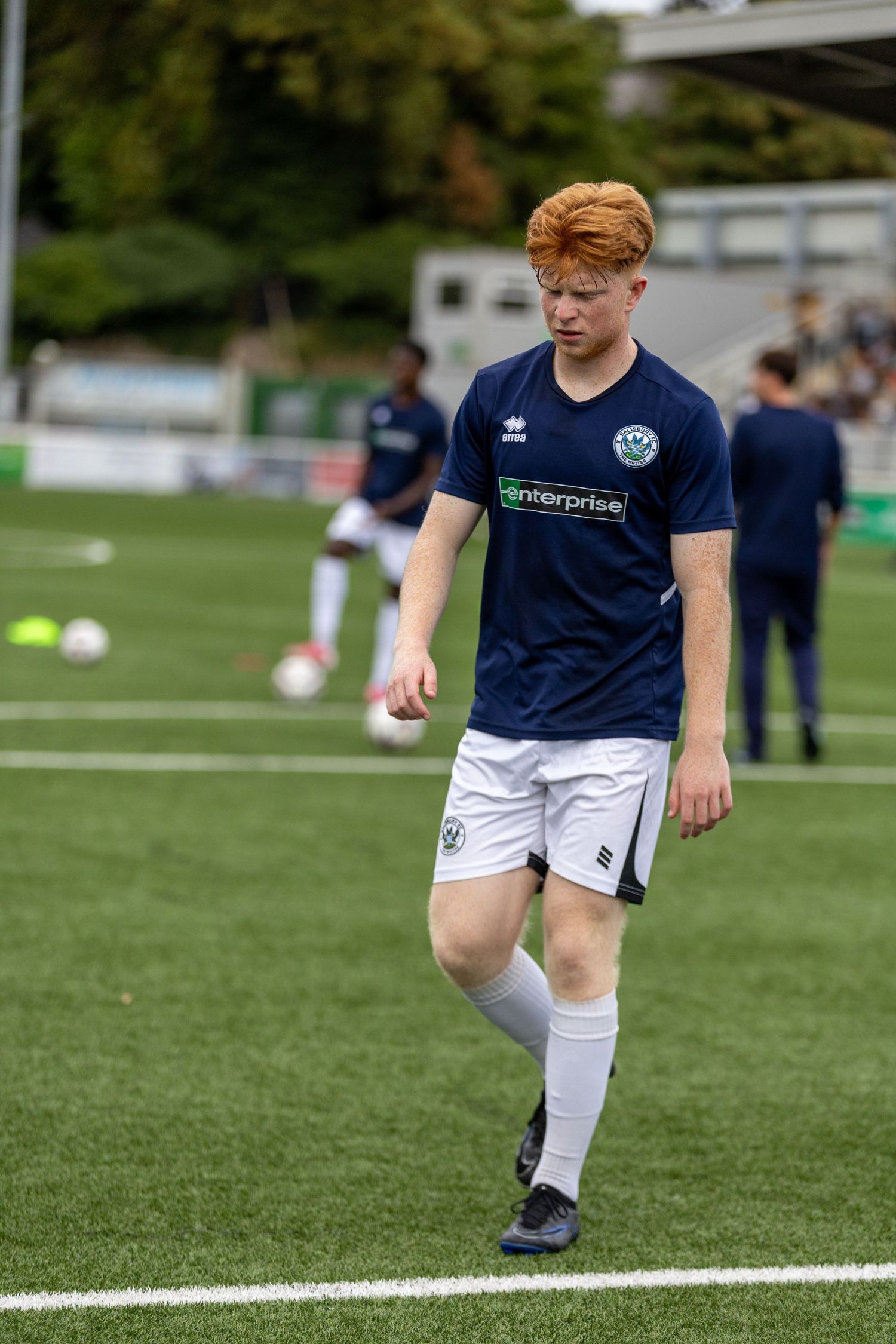 Young male soccer player in navy blue kit with orange hair walking on green turf during a football match warm-up.