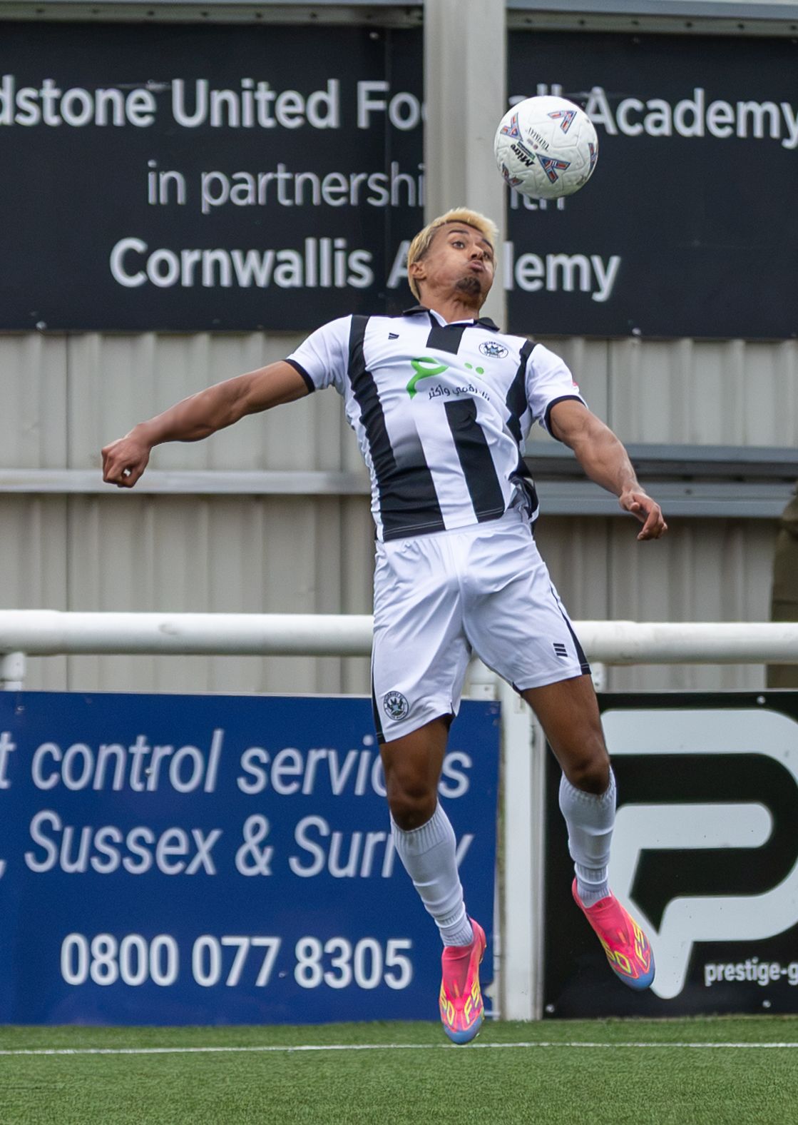 A footballer in black and white kit jumps to head the ball during a match at Maidstone United Football Academy.