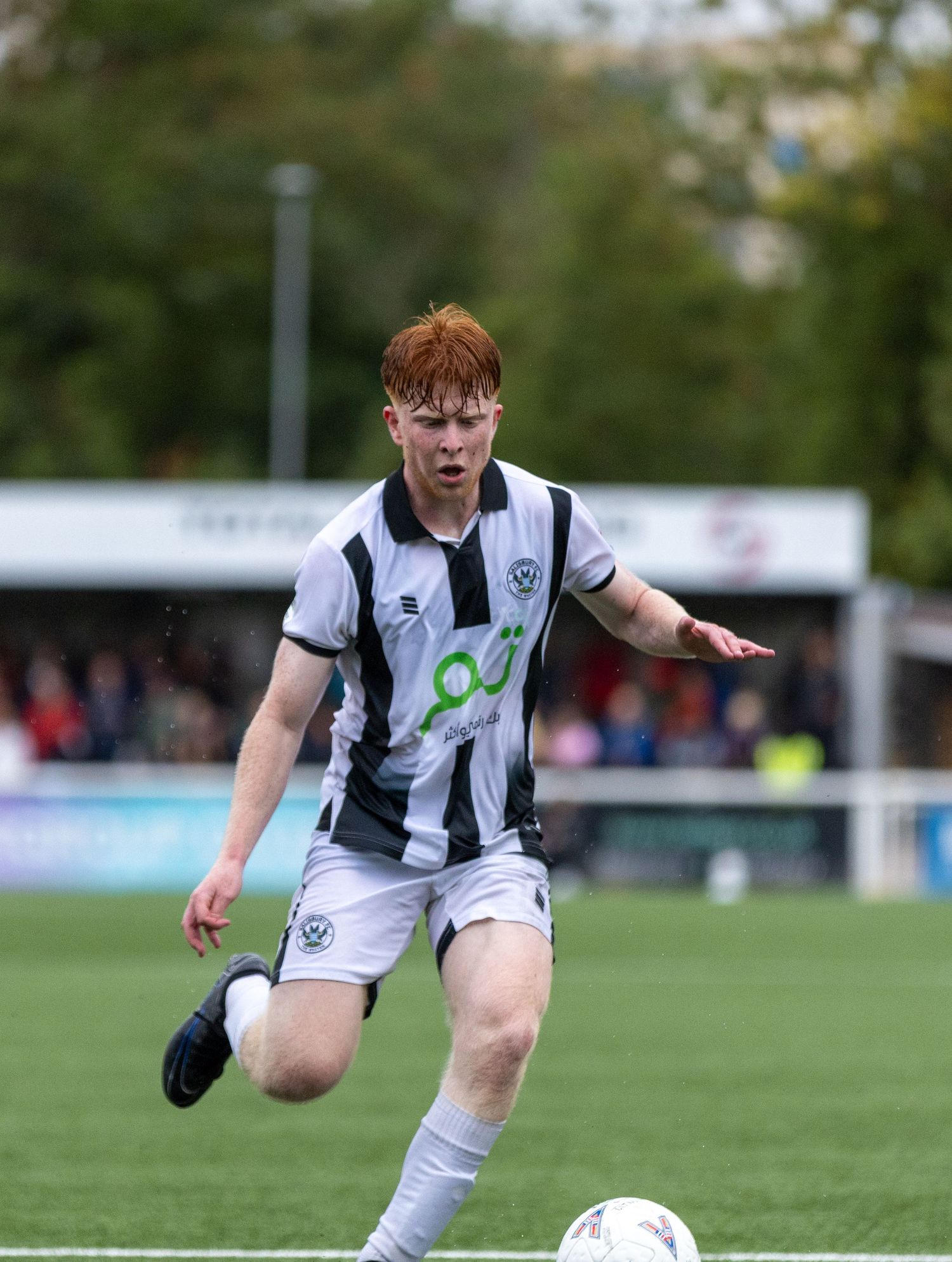 Young male soccer player in black and white striped kit dribbling a ball on a green pitch during a match.