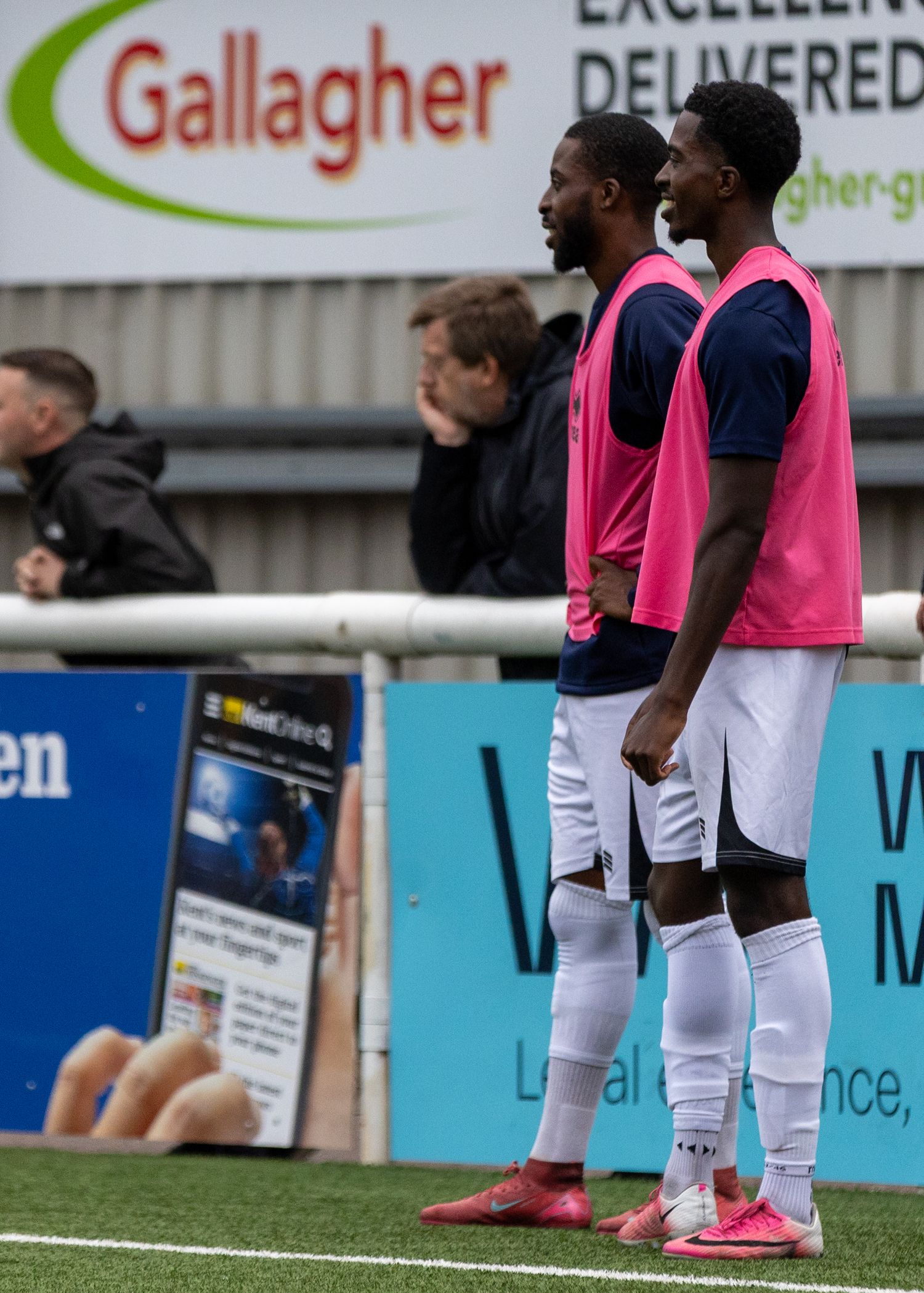 Two soccer players in pink bibs stand on the sideline near Gallagher advertising boards during a match.