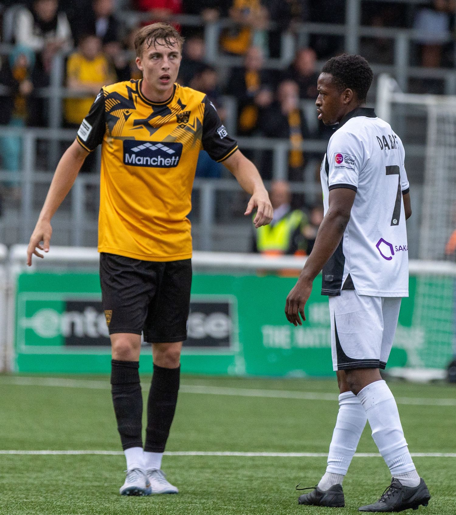 Two soccer players in a tense moment during a match, one in yellow-black and one in white, on a green pitch.