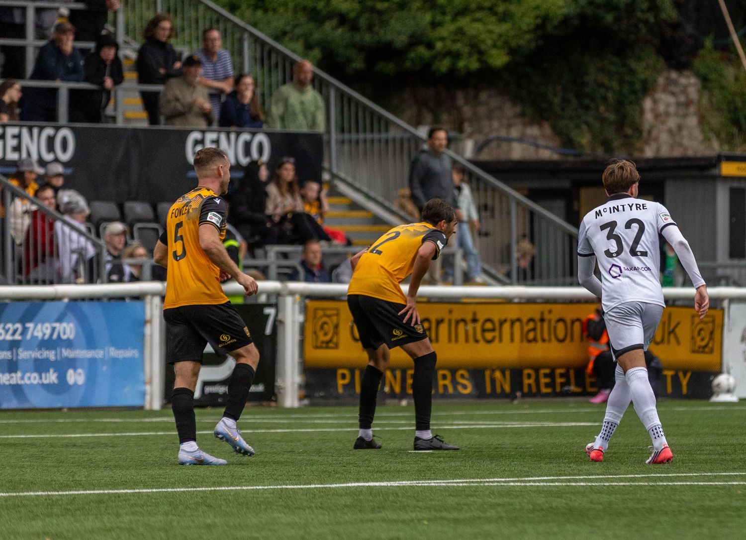 Soccer players in yellow and black uniforms compete on a green pitch during a match with spectators in the stands.