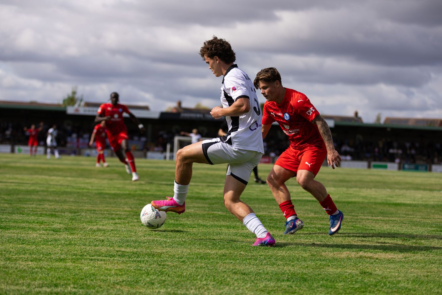 Two soccer players compete for the ball during an outdoor match, one in white and one in red, on a green pitch under cloudy skies.
