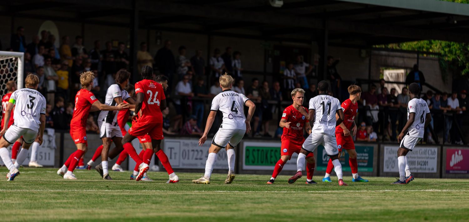 Soccer players in red and white uniforms compete for the ball during an outdoor match on a sunny day.