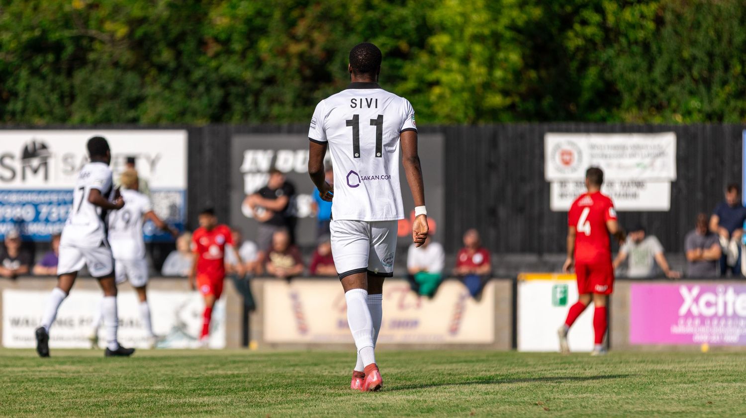 Soccer player wearing number 11 jersey walks on field during a sunny outdoor match with red and white teams.