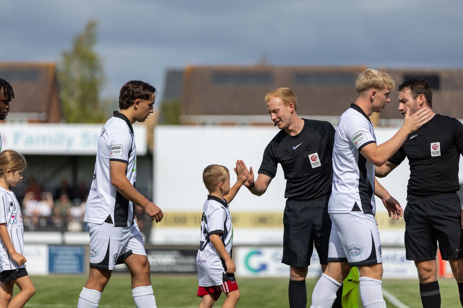 Soccer players shake hands with referee and mascot before a match on a sunny day at a local football ground.