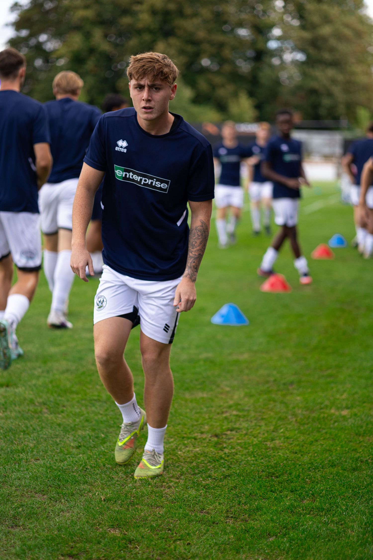 Young footballer in navy Enterprise-sponsored kit runs through training cones on grass pitch during team warm-up session.