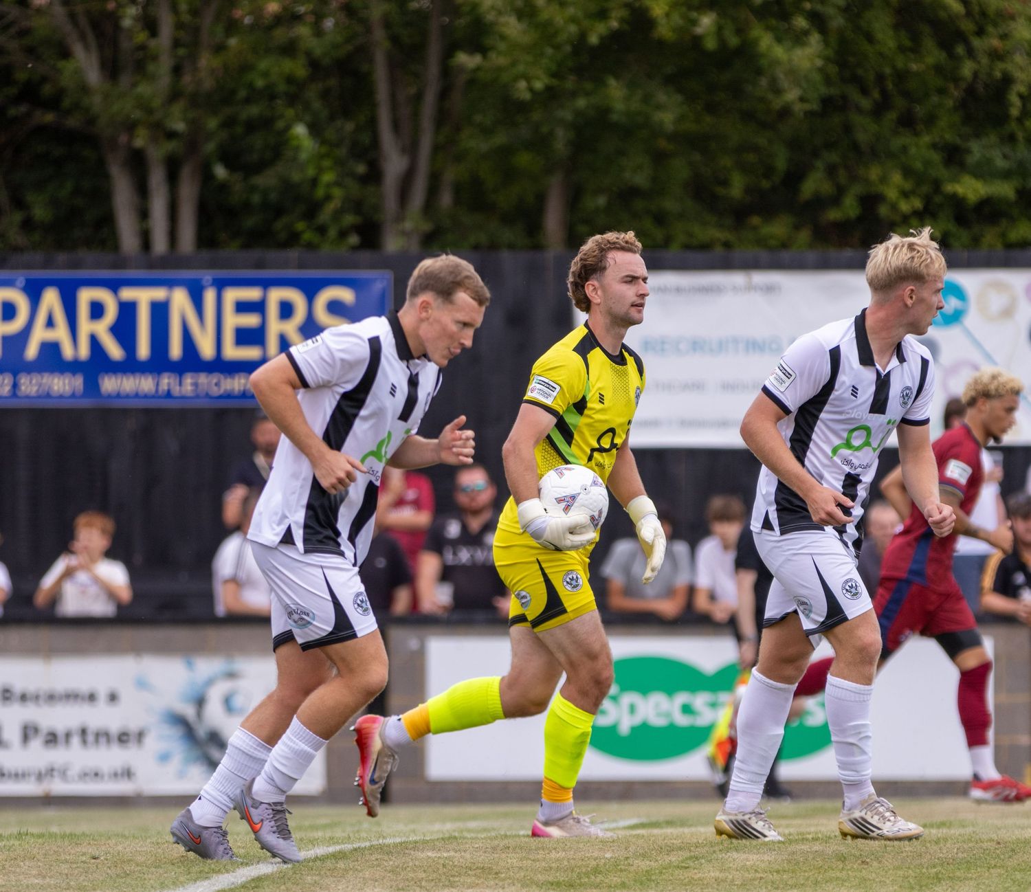 Football players in black and white kit compete with a yellow goalkeeper during a non-league soccer match.