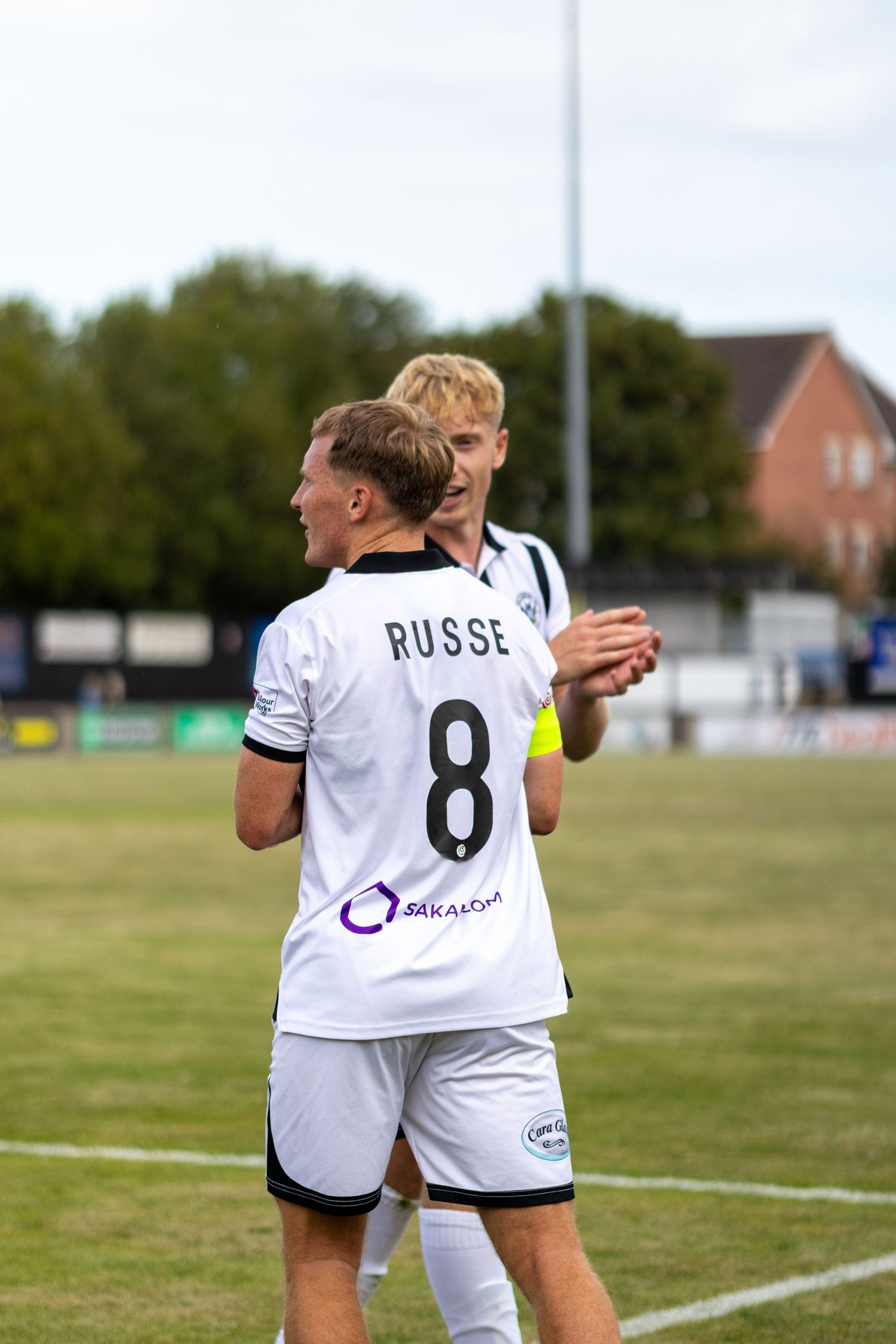 Two football players in white jerseys celebrate on the pitch, one wearing number 8 RUSSE shirt with SAKAN sponsor.
