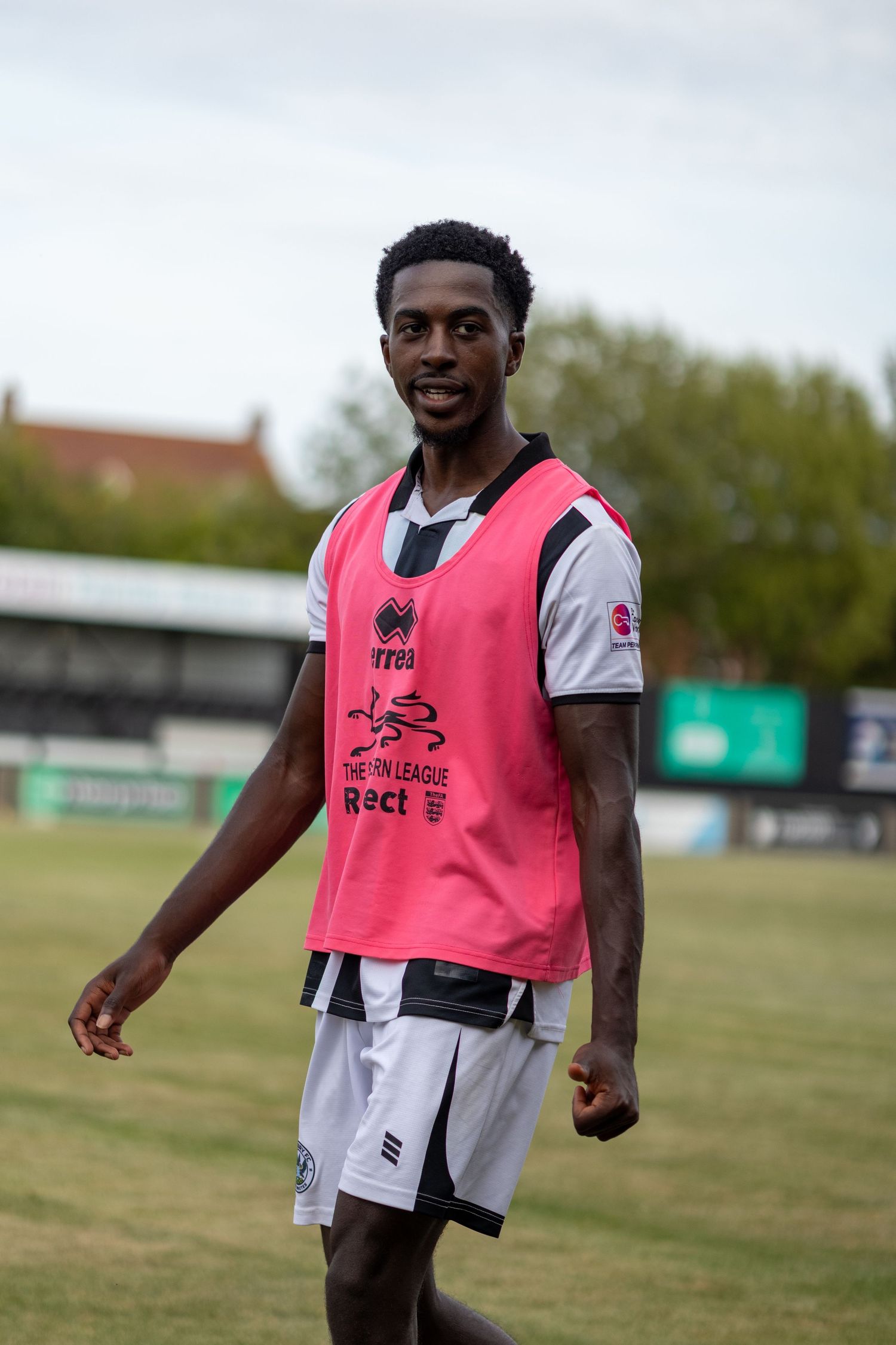 A male soccer player in a pink training bib over a white and black kit smiles on a grass field.