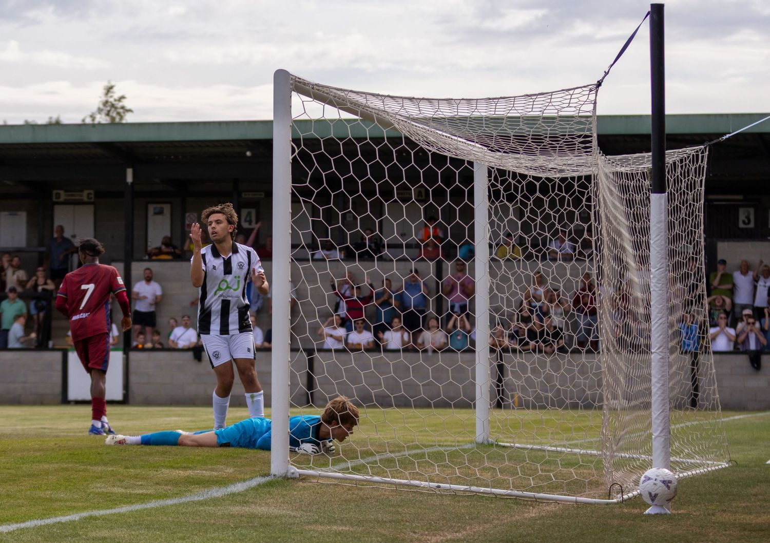 Football player in black and white stands by the goal as goalkeeper lies on the ground after conceding.