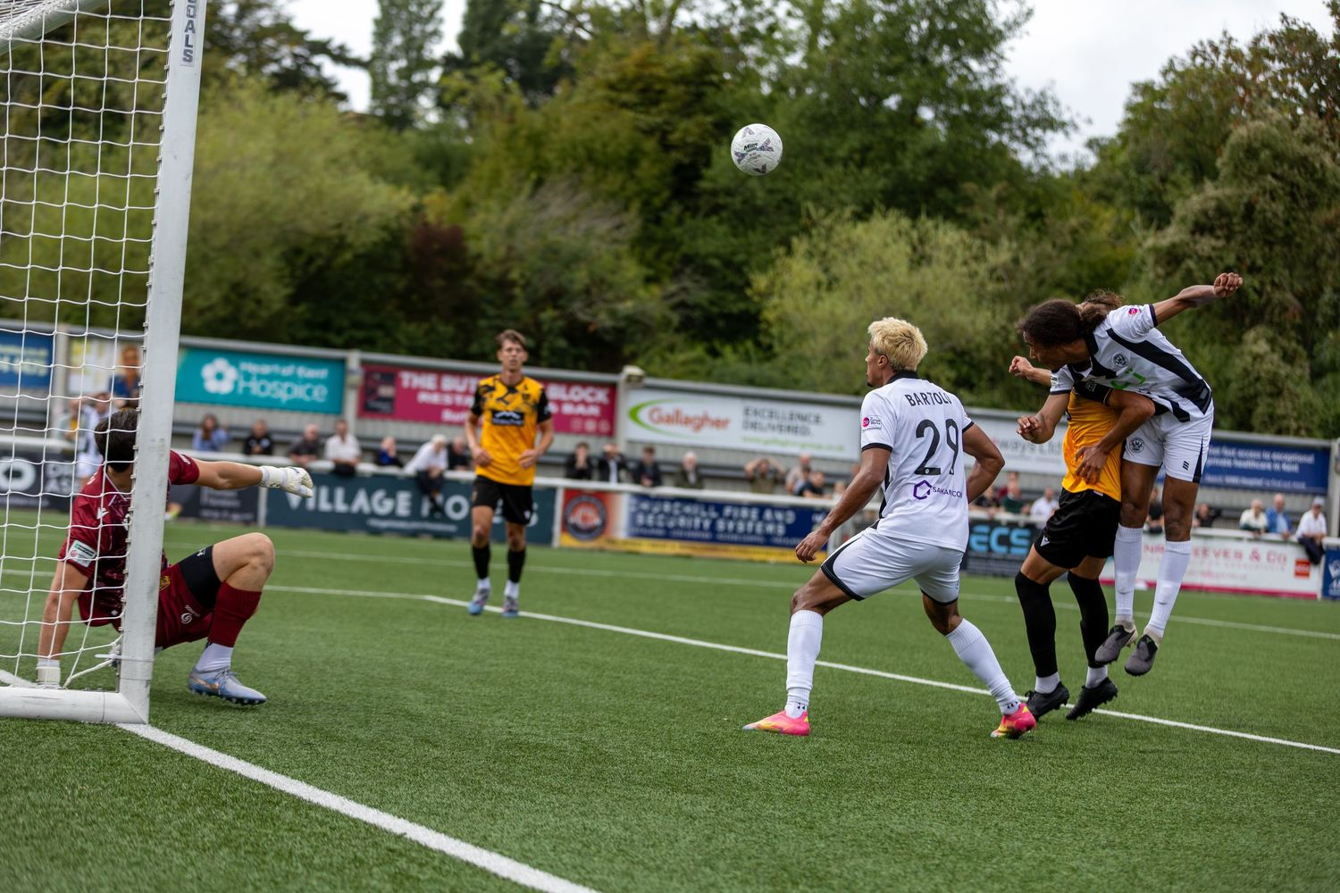Soccer players compete in a header duel near the goal during an outdoor match on a green pitch.