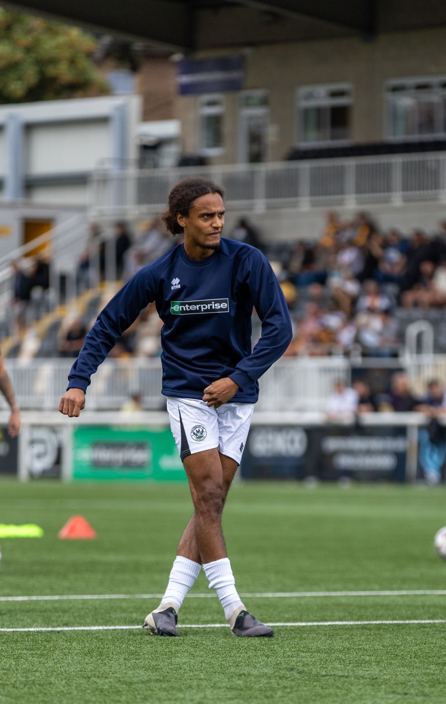 Soccer player in navy Enterprise-sponsored training top and white shorts warming up on a green pitch before a match.