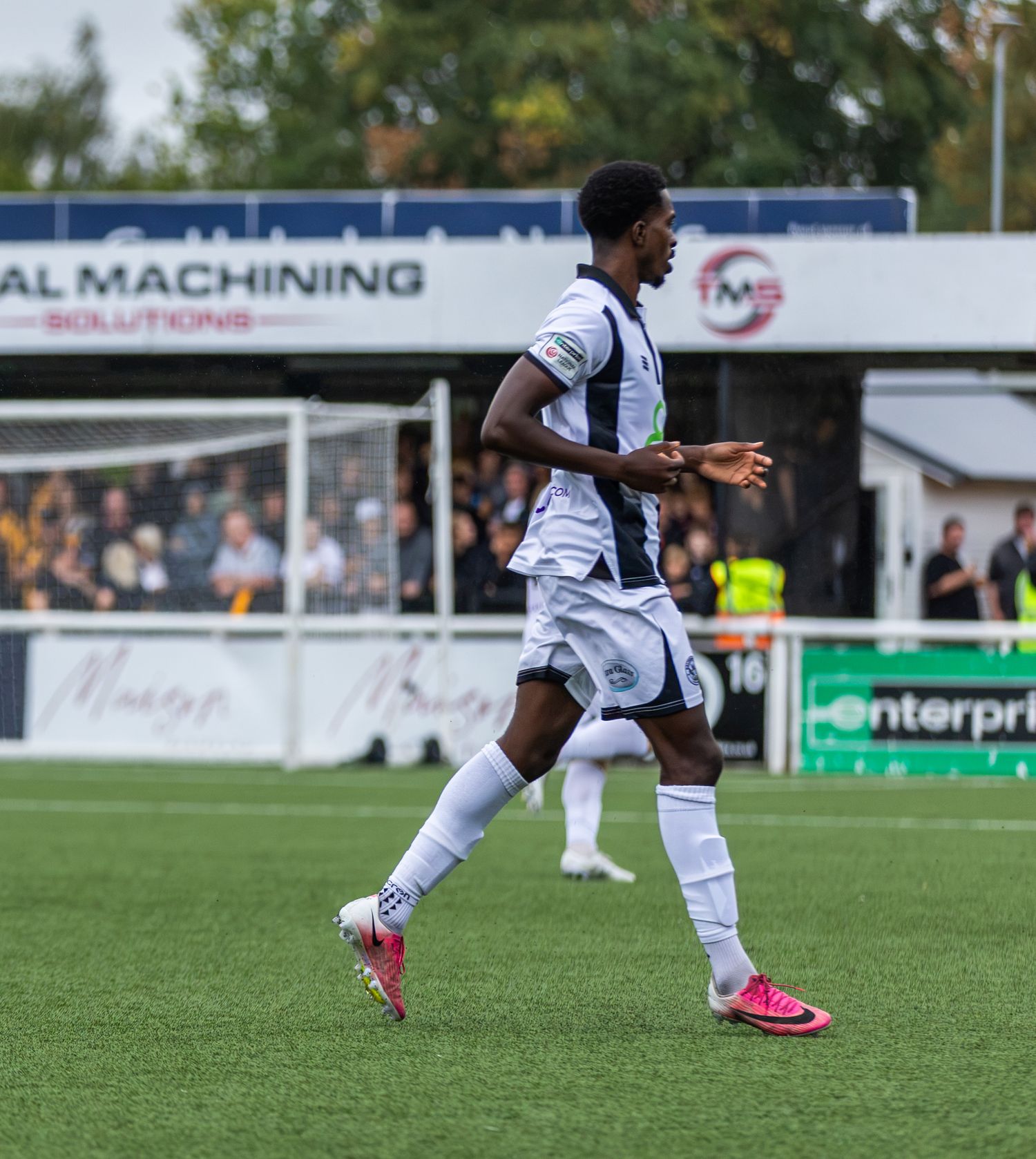 A football player in white kit runs on a green pitch during a match at a non-league ground.