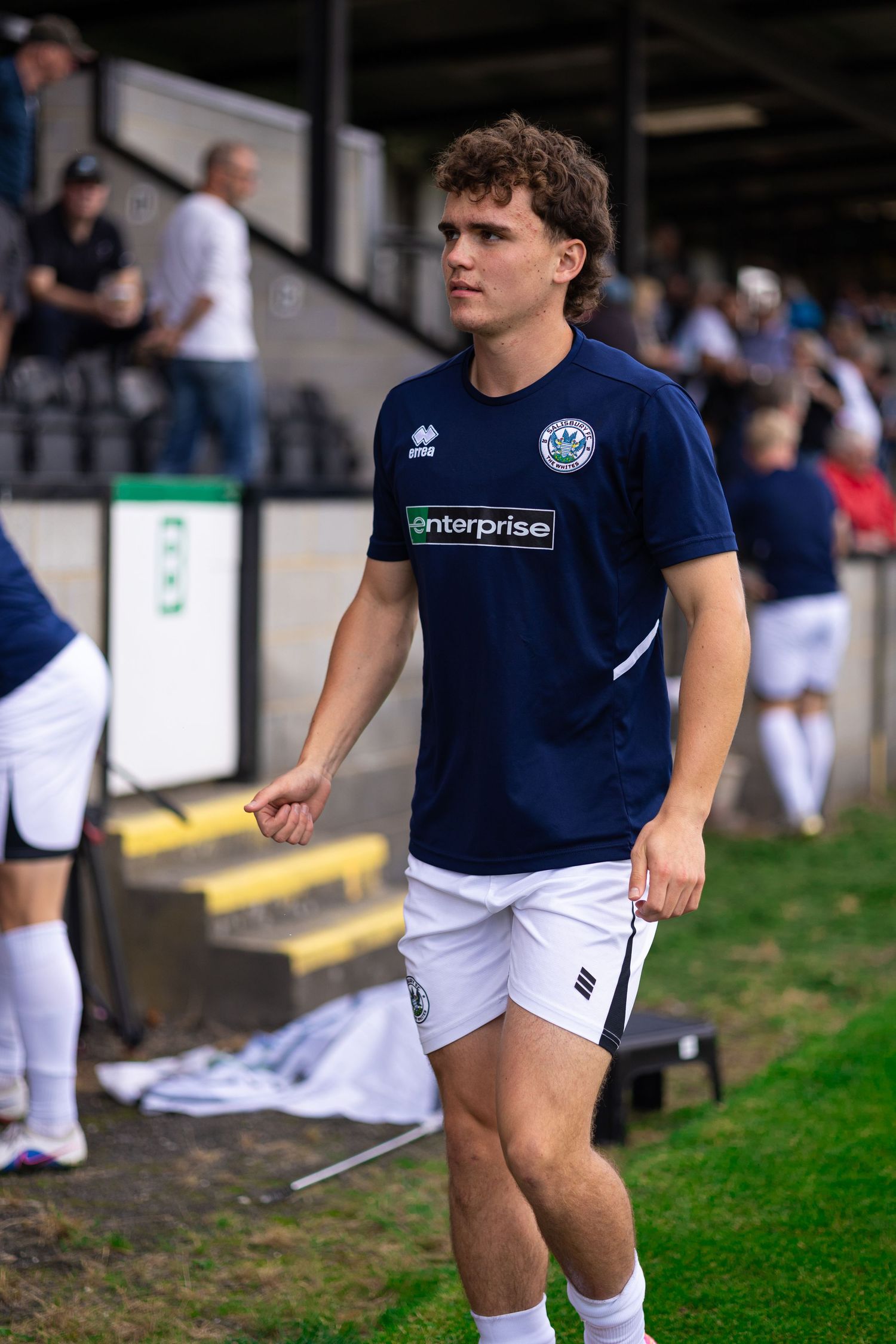 Young footballer in navy Enterprise-sponsored jersey and white shorts walks near the sideline during a match.