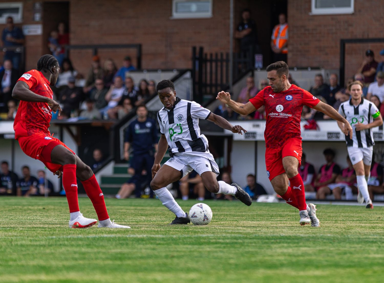 Football players in black-white and red kits compete for the ball during an outdoor match on a grass pitch.