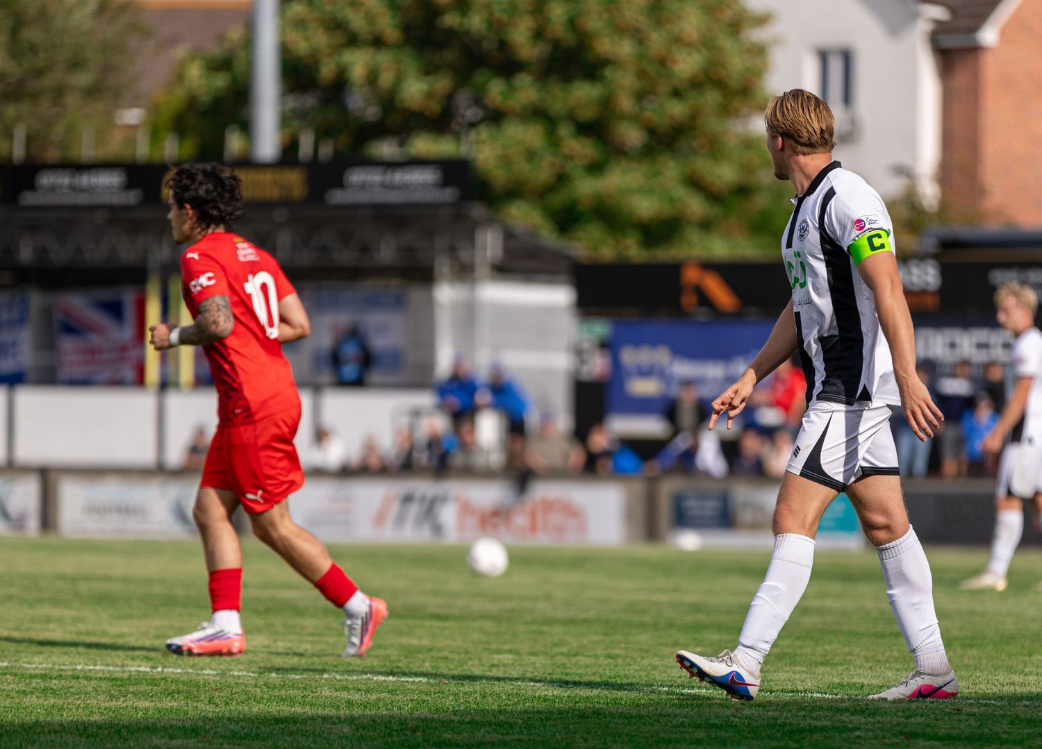 Two soccer players in red and black-and-white uniforms compete on a green pitch during a sunny outdoor match.