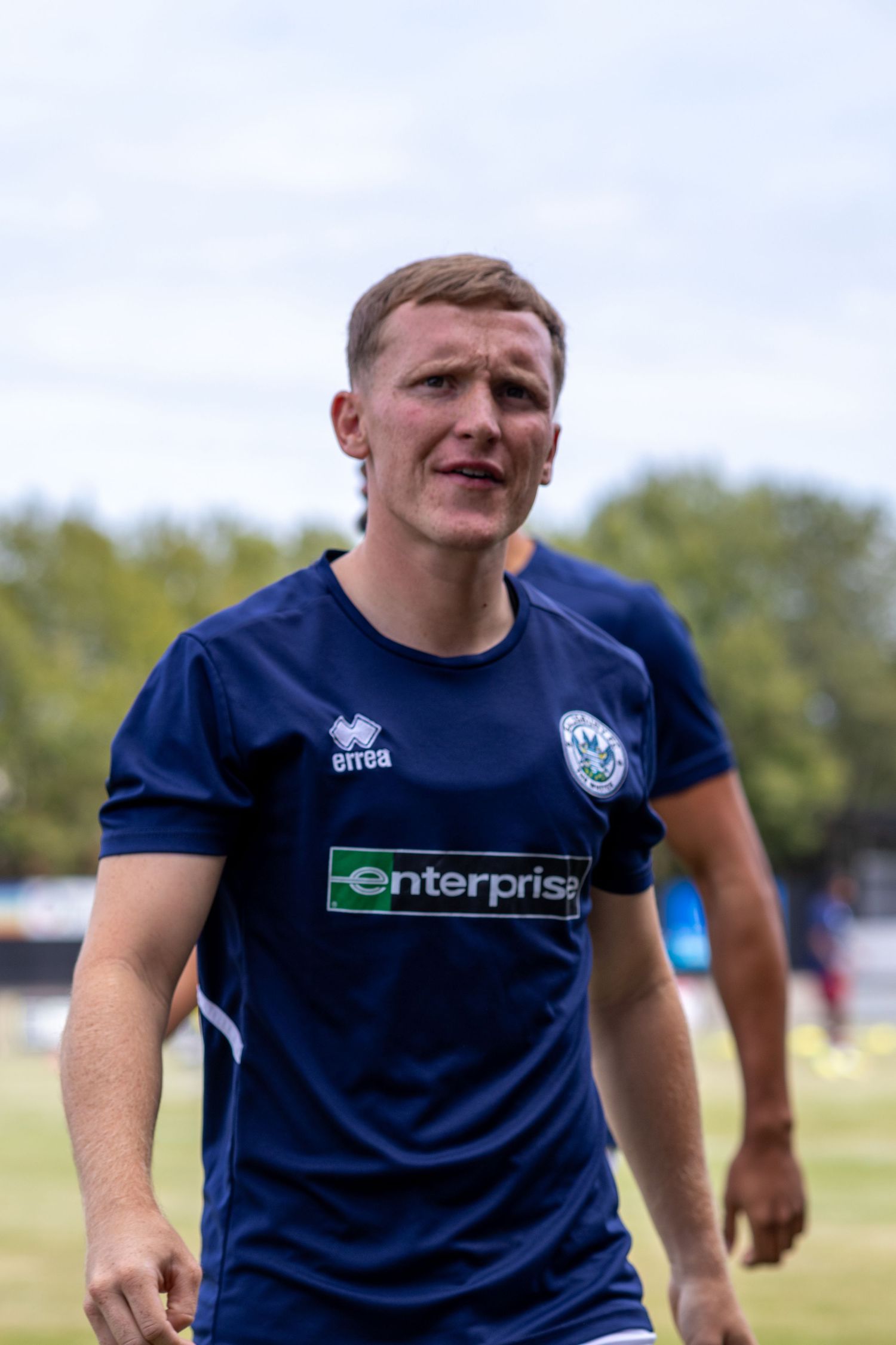 Male footballer in navy Errea Enterprise-sponsored shirt walking across football pitch during warmup.