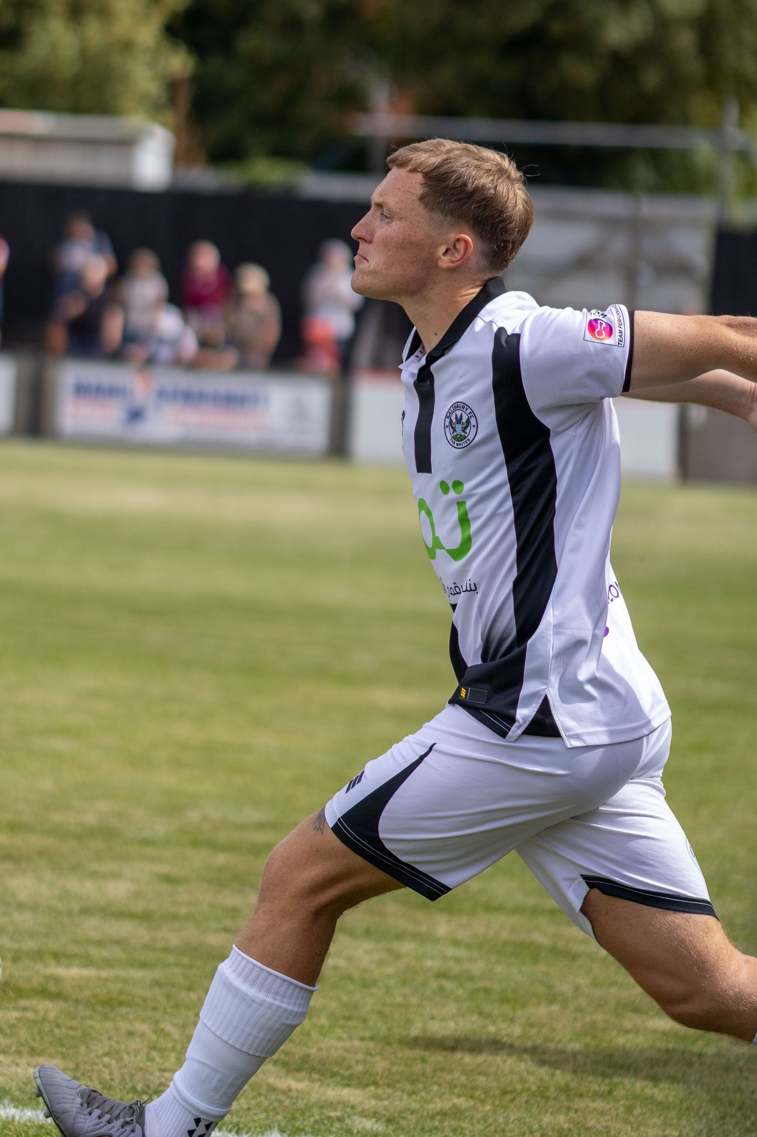 A blonde male footballer in a white and black kit strides across a grass pitch during a sunny afternoon match.