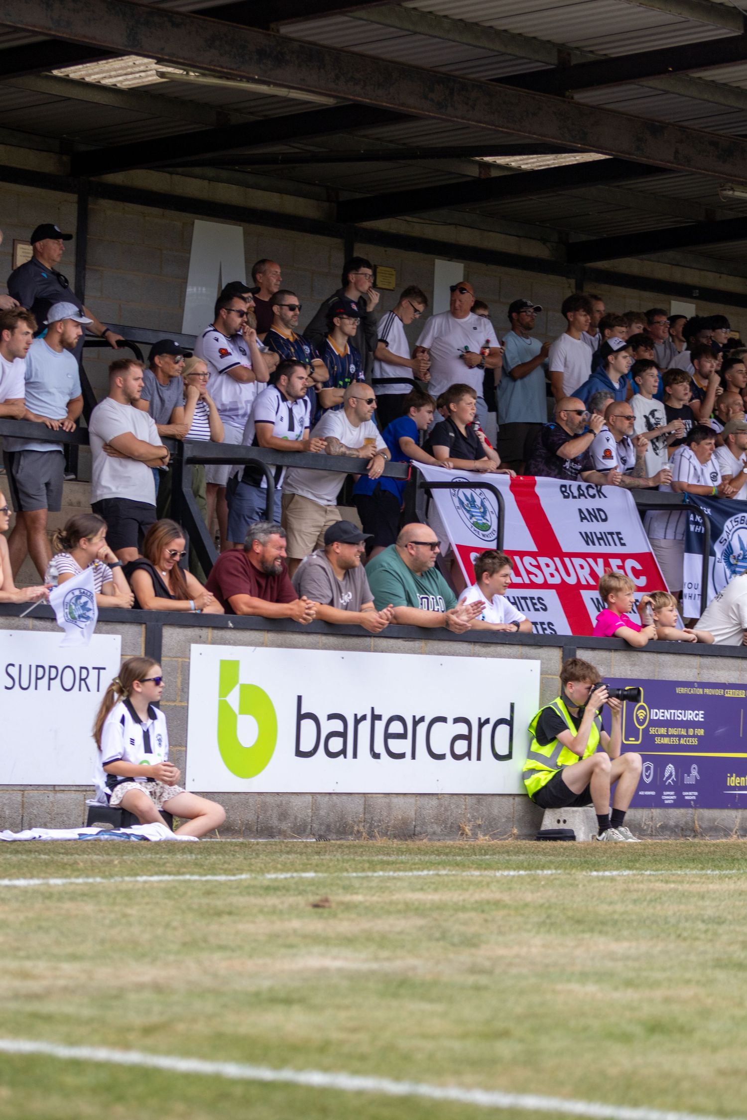 Large crowd of Salisbury FC fans watching from the stands with club banner on a sunny matchday.