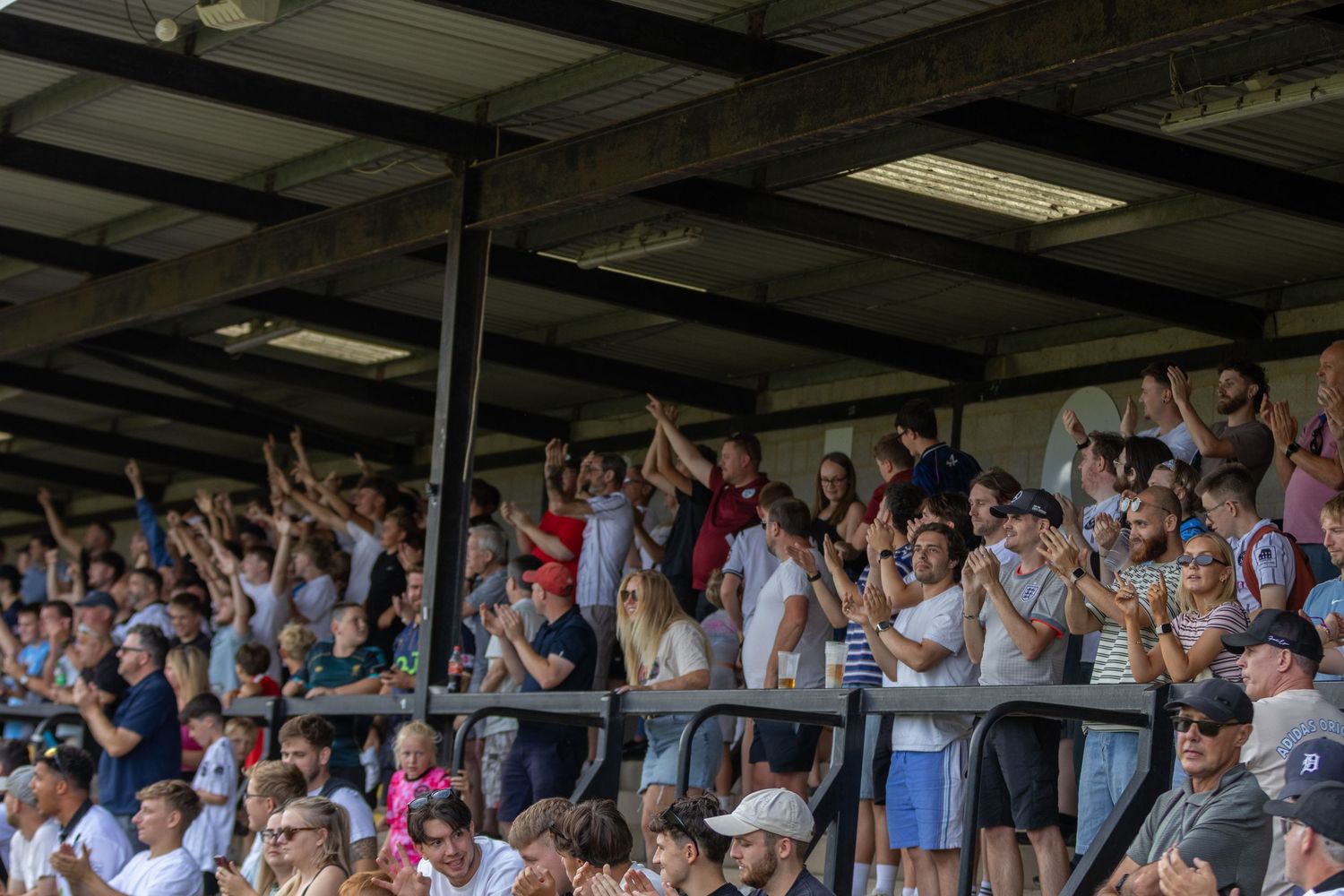 Excited football fans cheer and celebrate in a covered stadium stand on a sunny matchday.