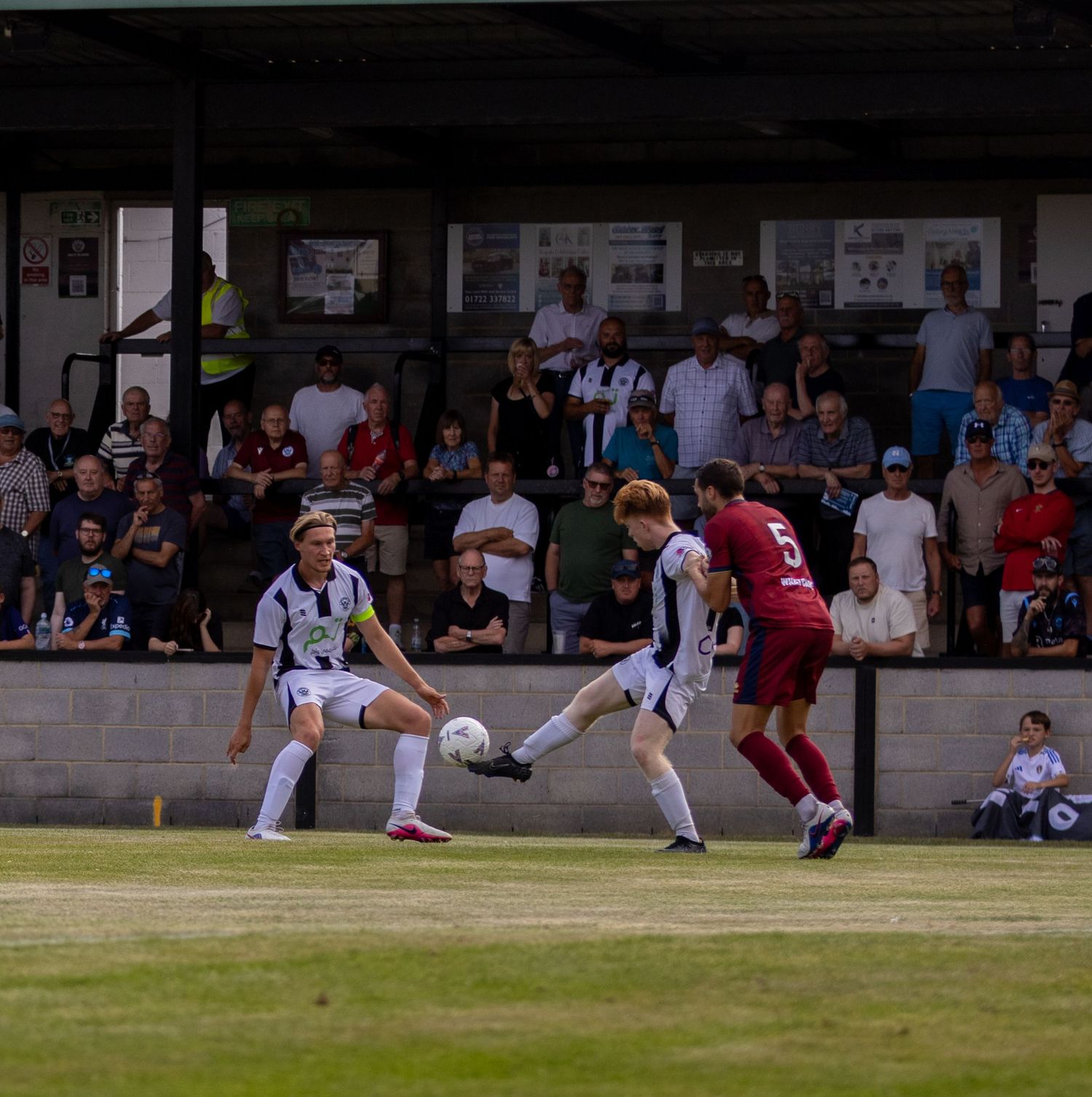 Football players compete for the ball near the touchline as spectators watch from the stands behind them.