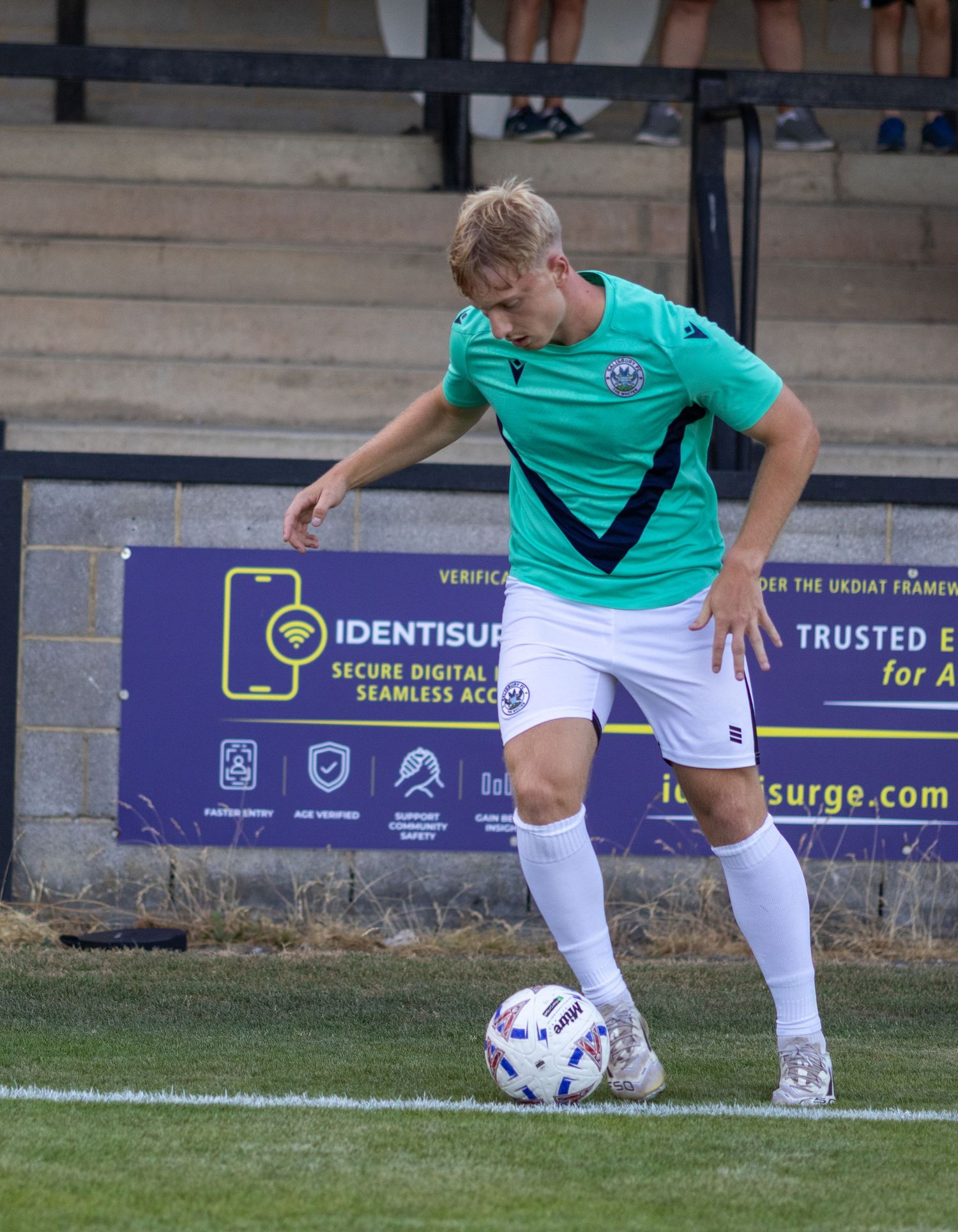 A soccer player in a teal kit controls the ball during a match near an Identisurge advertising board.