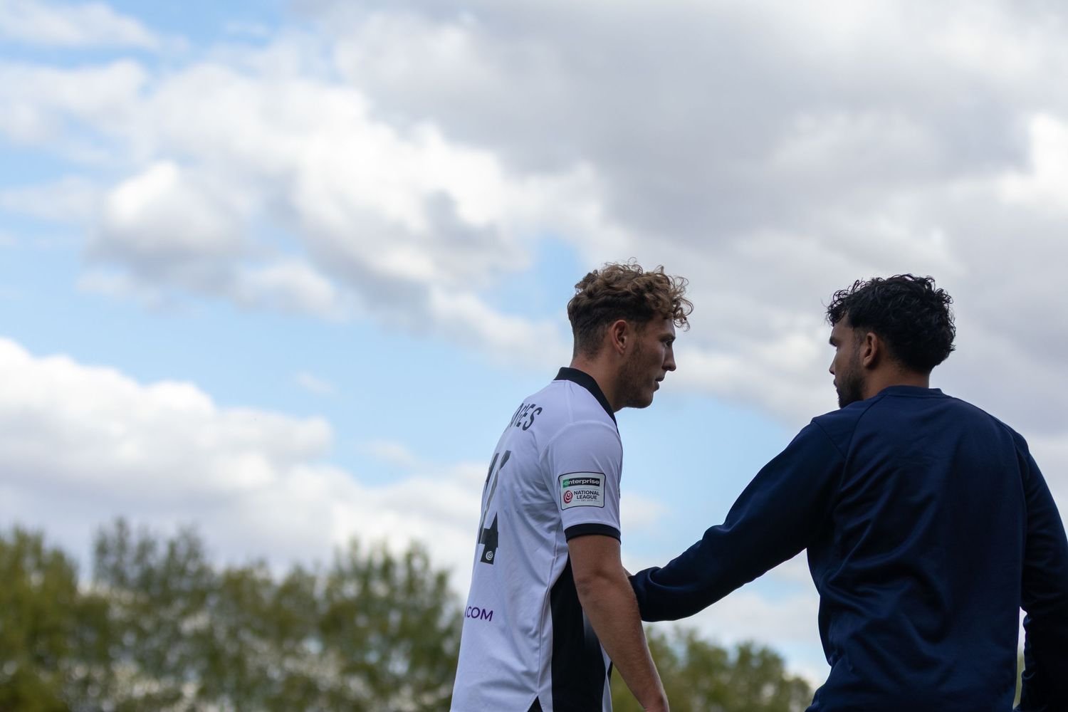 Two male soccer players in conversation on a field, one in white jersey, under a cloudy sky.