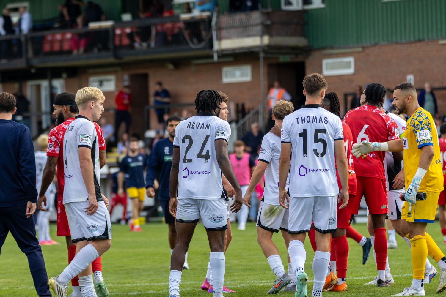 Soccer players in white and red jerseys walk on a green pitch during a competitive match at a stadium.
