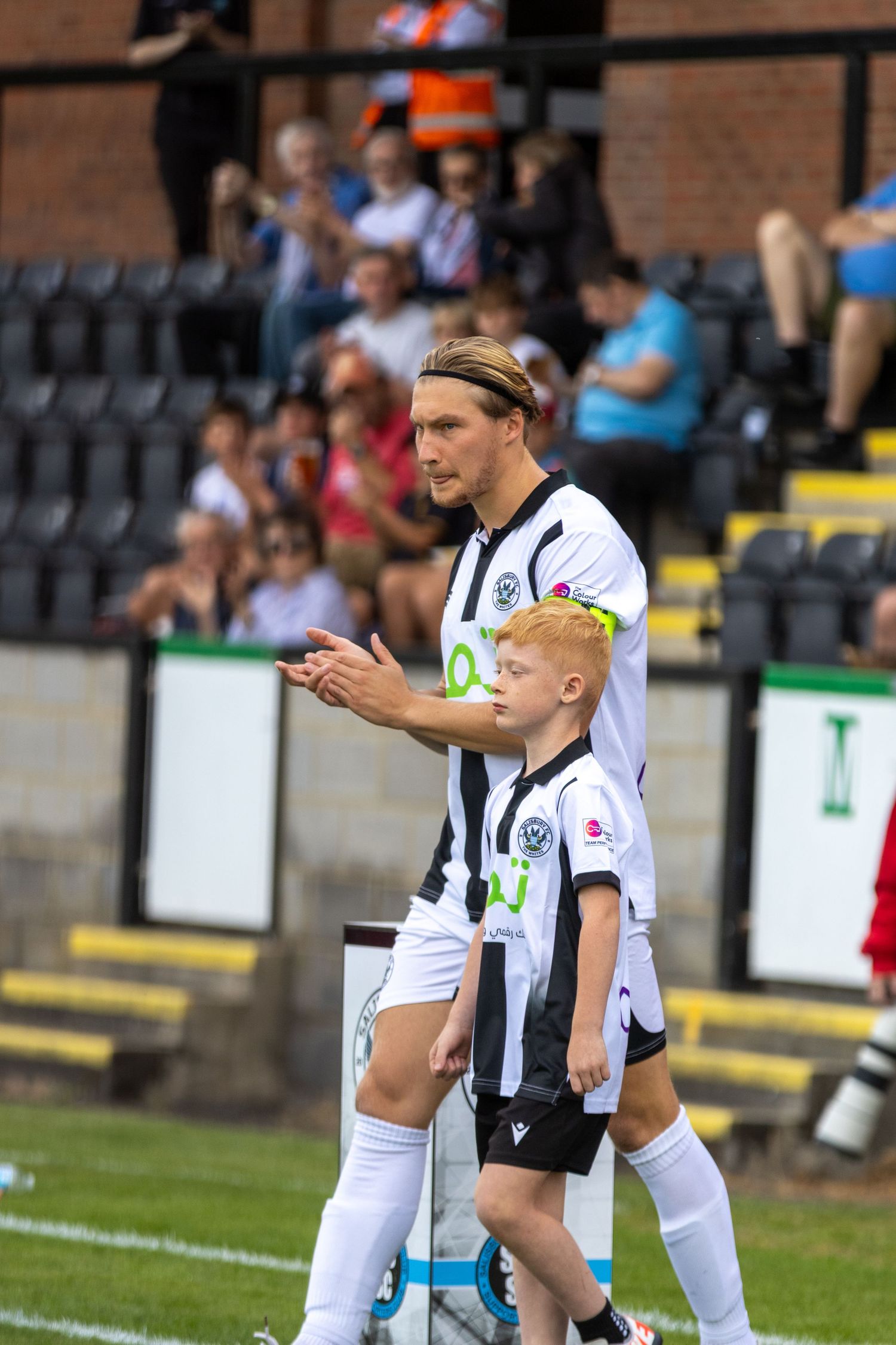 A footballer in black and white striped kit walks onto the pitch with a young mascot beside him.