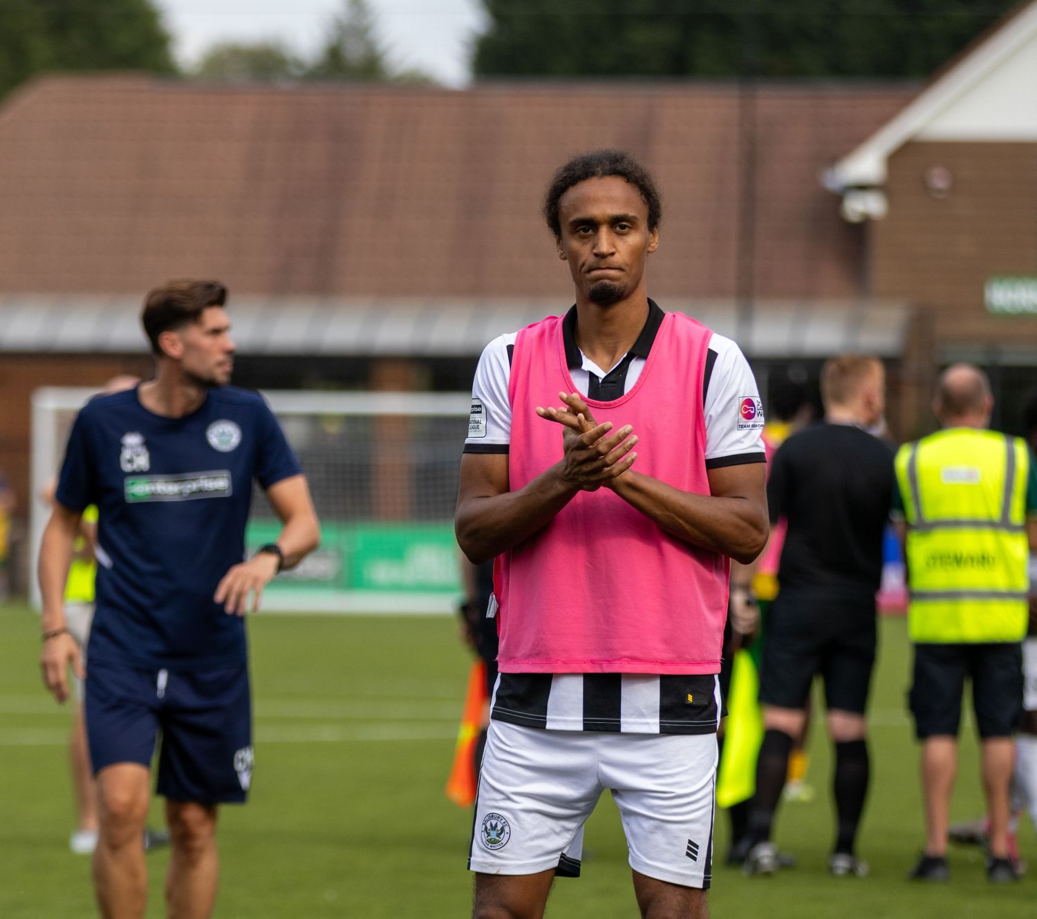 A footballer wearing a pink bib over black and white kit applauds after a match on a green pitch.