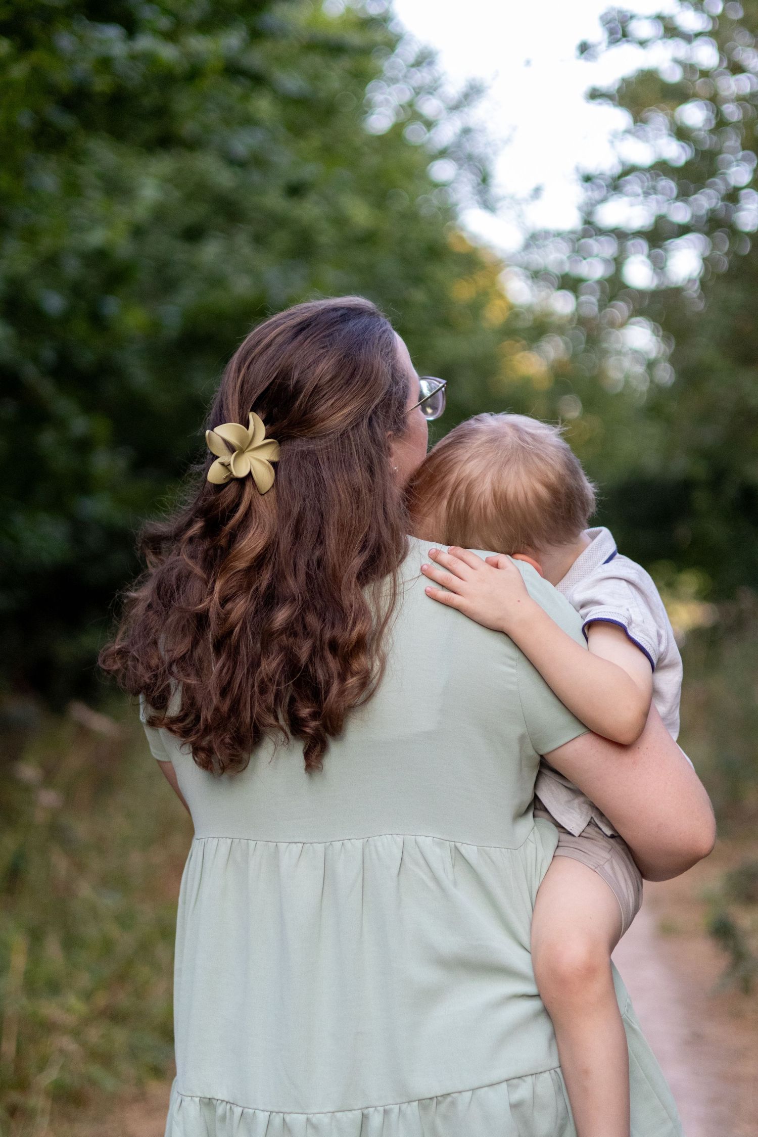 A mother in a mint tiered dress holds her sleepy baby against her shoulder on a tree-lined trail.
